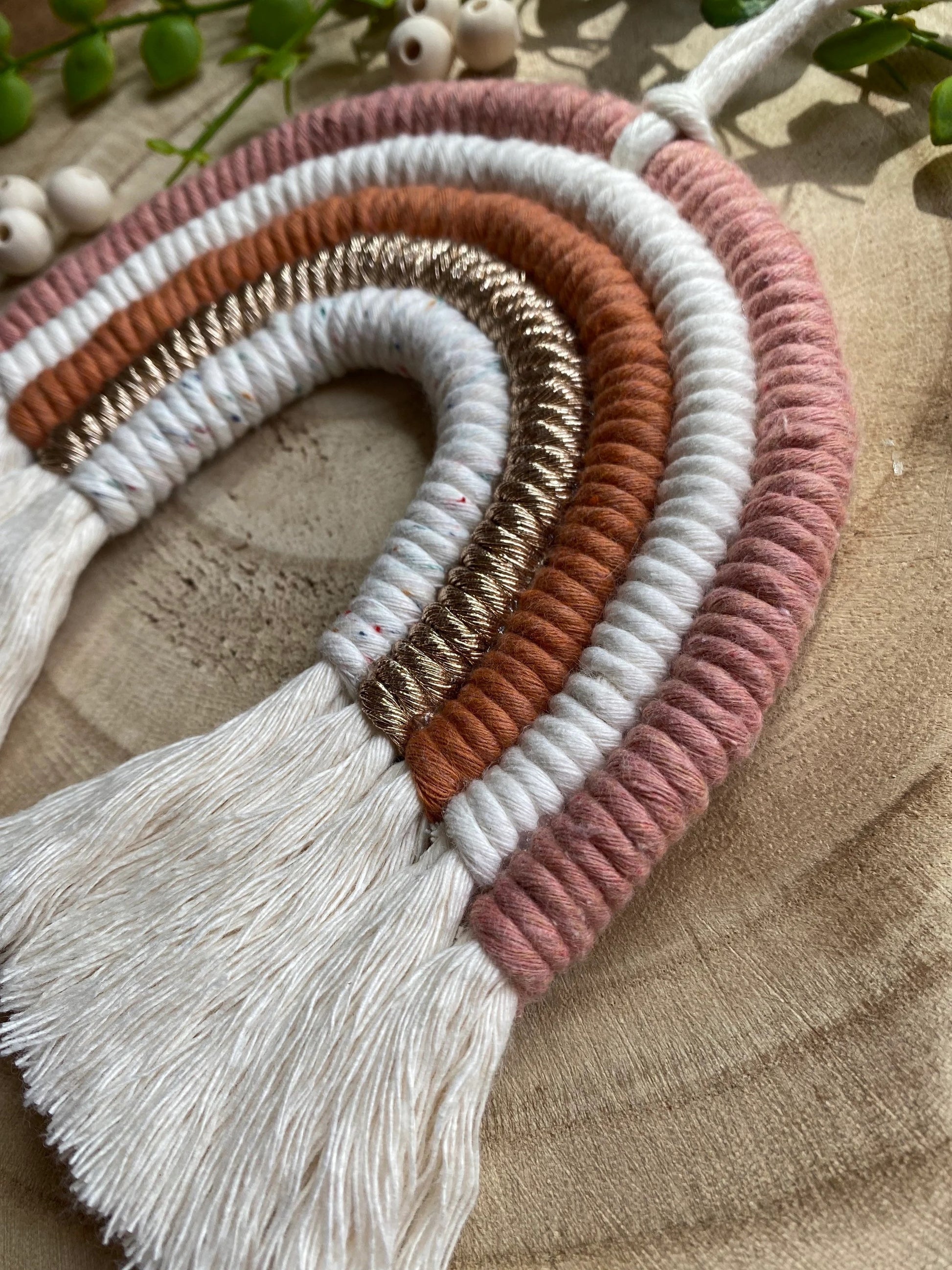 Close up of a handmade macrame rainbow wall hanging in shades of pink, white, and gold, with tassels on the ends. Displayed on a wooden board.