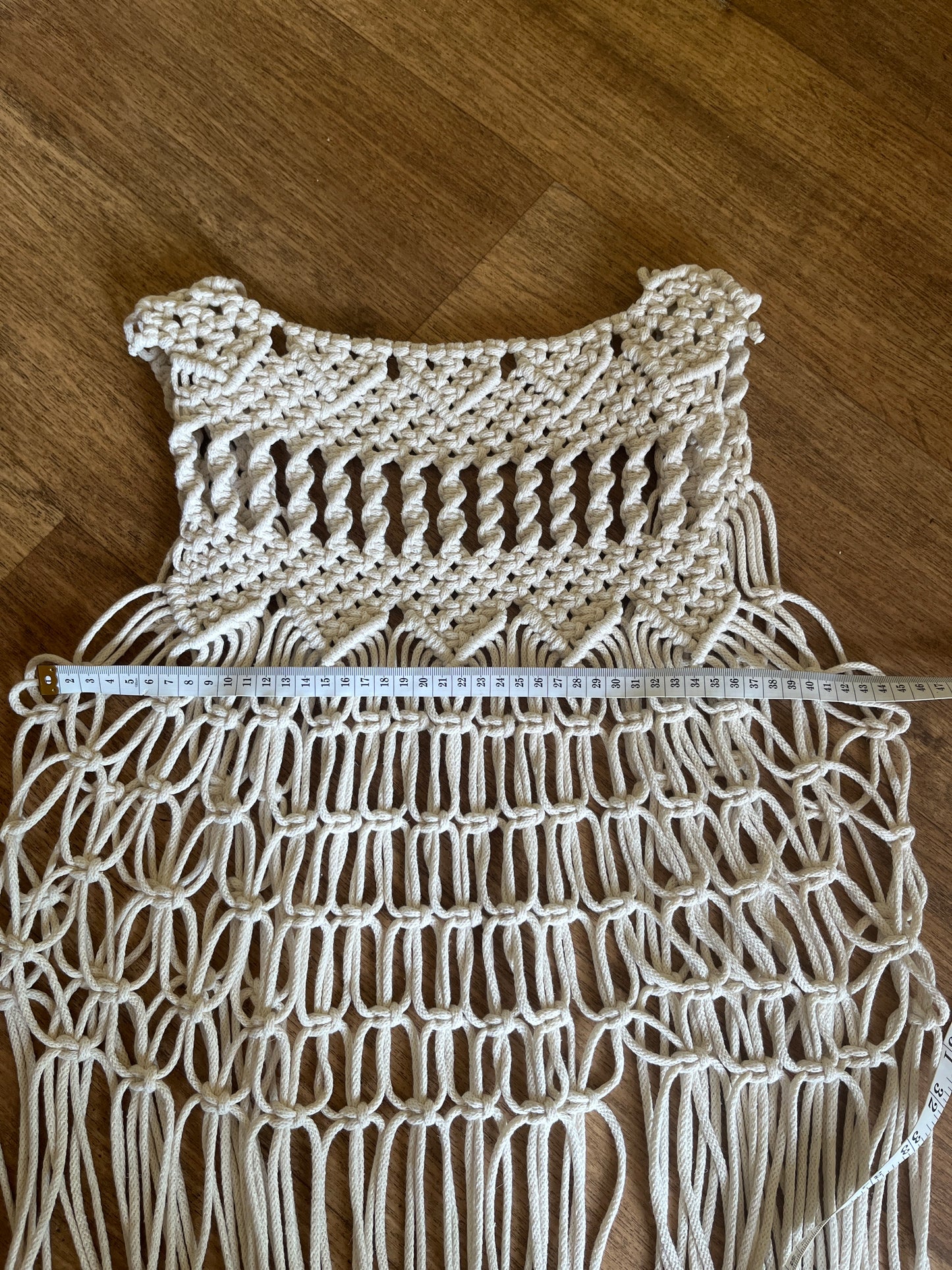 Close up of the backside of a handmade natural boho fringed waistcoat made with intricate macrame knots, laid flat on a wooden floor with a white tape measure laid horizontally across it.