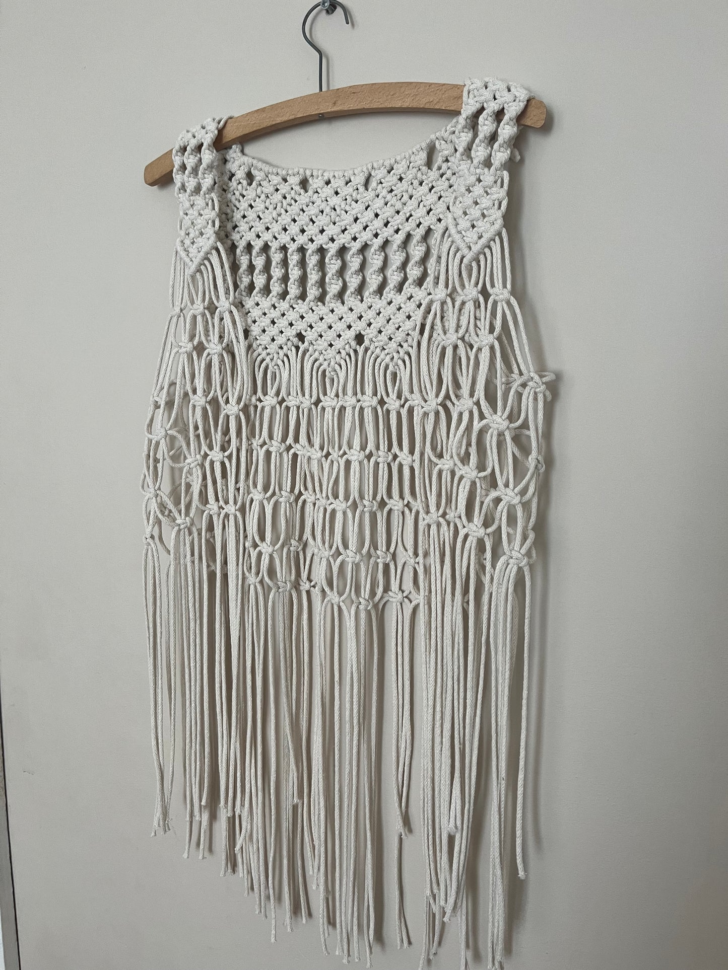 A handmade natural boho fringed waistcoat made with intricate macrame knots, hanging on a wooden coat hanger against a beige wall.