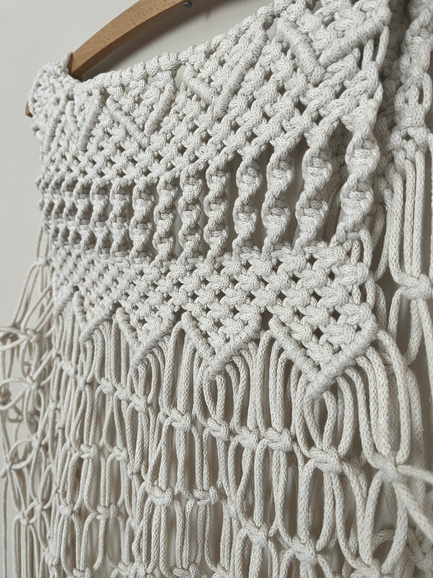 Close up of a handmade natural boho fringed waistcoat made with intricate macrame knots, hanging on a wooden coat hanger against a beige wall.