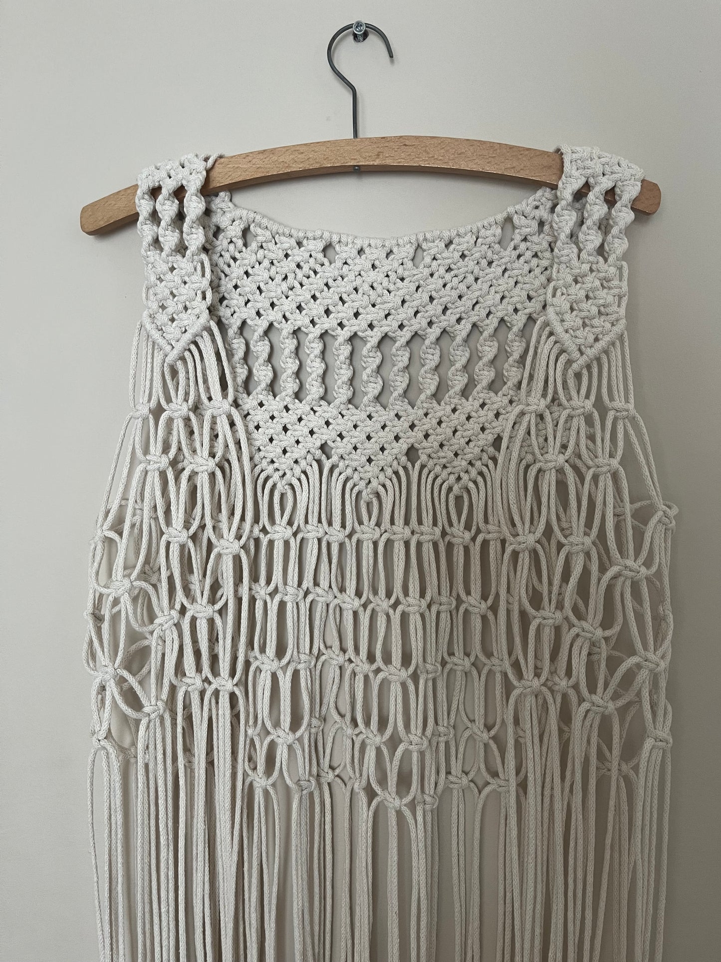 A handmade natural boho fringed waistcoat made with intricate macrame knots, hanging on a wooden coat hanger against a beige wall.