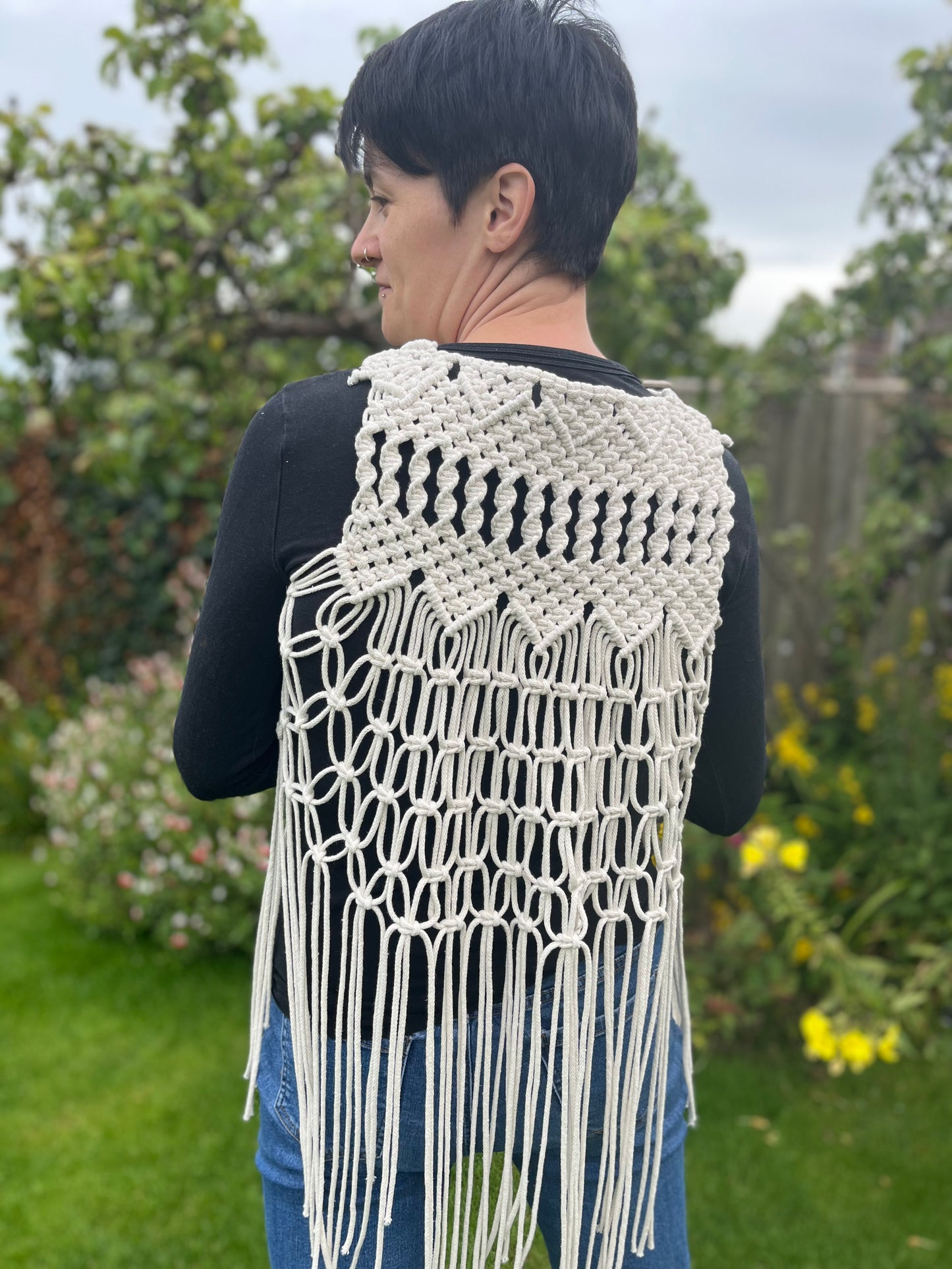 A person with short brown hair stands outside with her back to the camera, she is wearing a natural boho fringed waistcoat over a black long sleeved top.