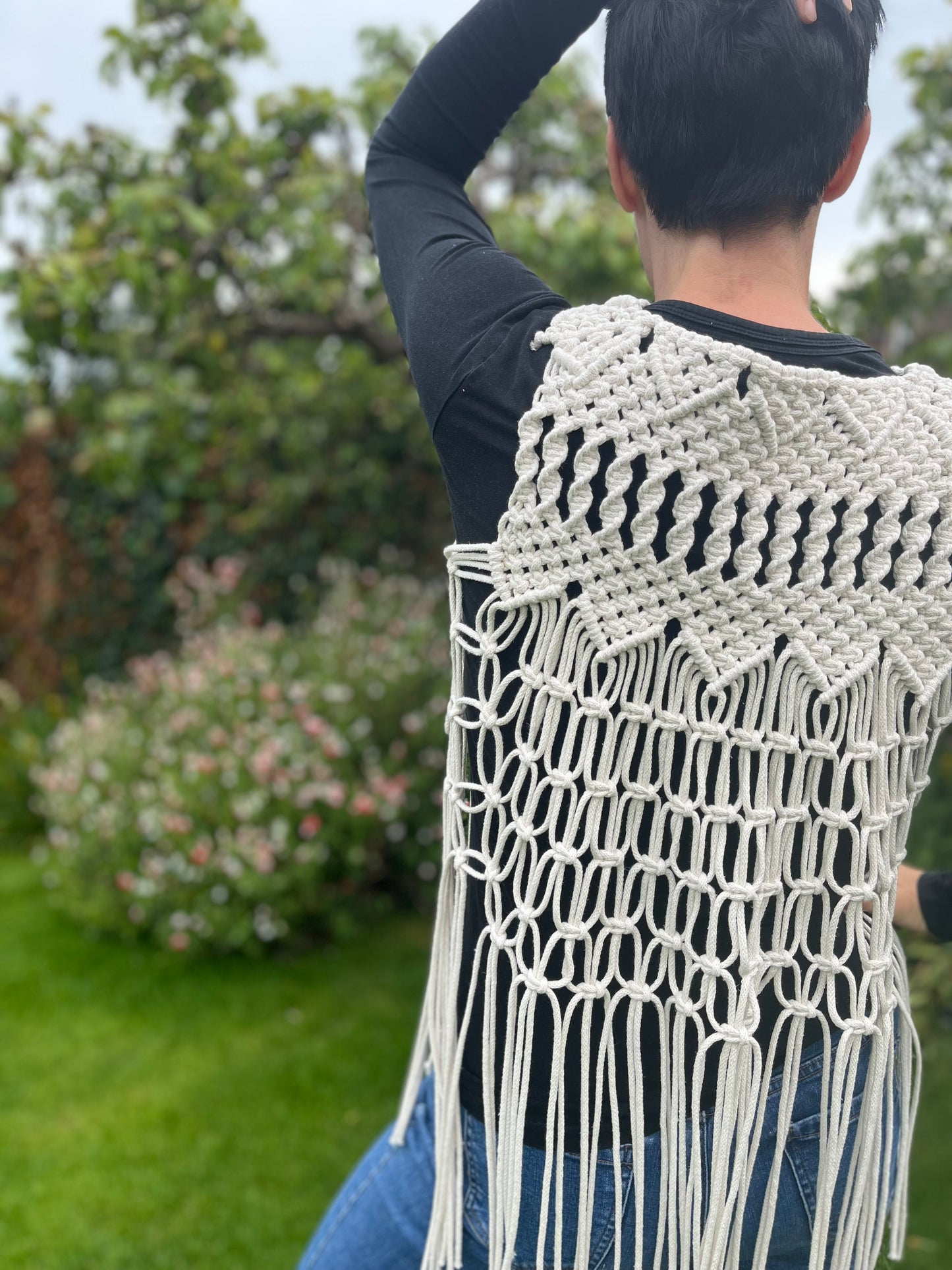 A person with short brown hair stands outside with her back to the camera, she is wearing a natural boho fringed waistcoat over a black long sleeved top, with a hand on the top of her head.