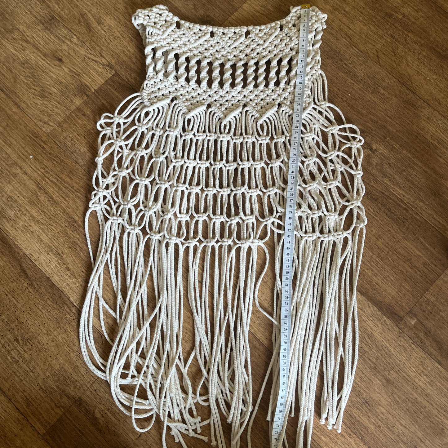 The reverse of a handmade natural boho fringed waistcoat made with intricate macrame knots, laid out flat on a wooden floor with a white tape measure vertically across it.