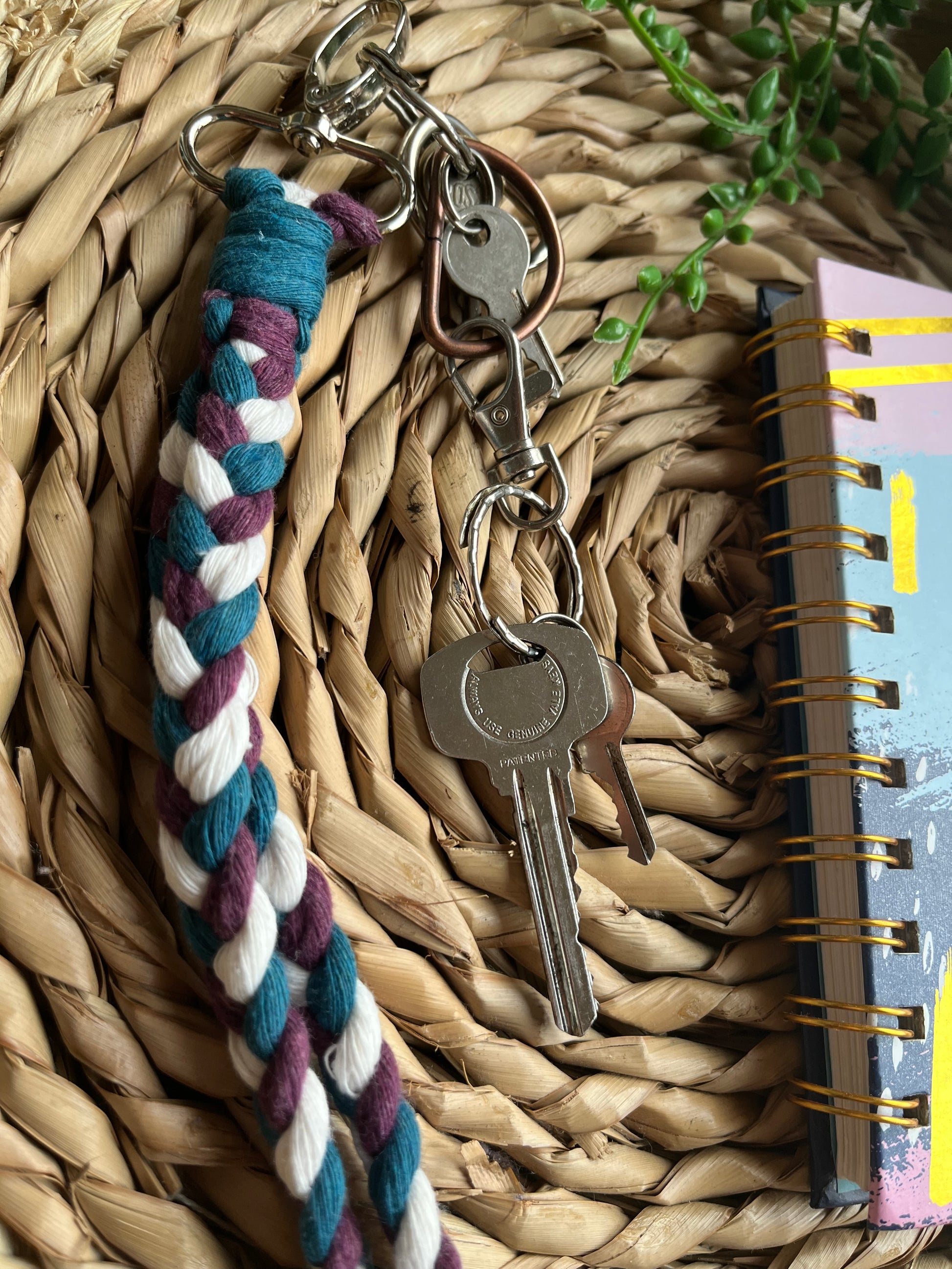 A photo of a handmade macrame braided wristlet attached to a small set of keys, laid on a wicker mat next to a notebook. The wristlet is made with petrol blue, berry purple and natural recycled cotton, with a metal lobster clasp.