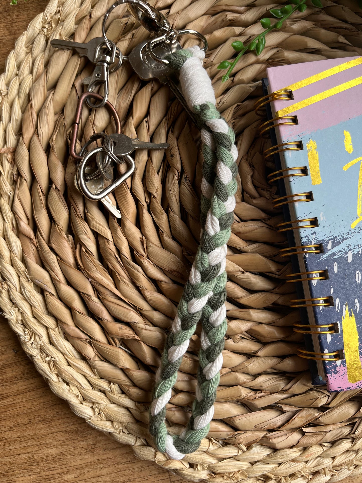 A photo of a handmade macrame braided wristlet attached to a smalls set of keys, laid on a wicker mat next to a notebook. The wristlet is made with green and natural recycled cotton, with a metal lobster clasp.