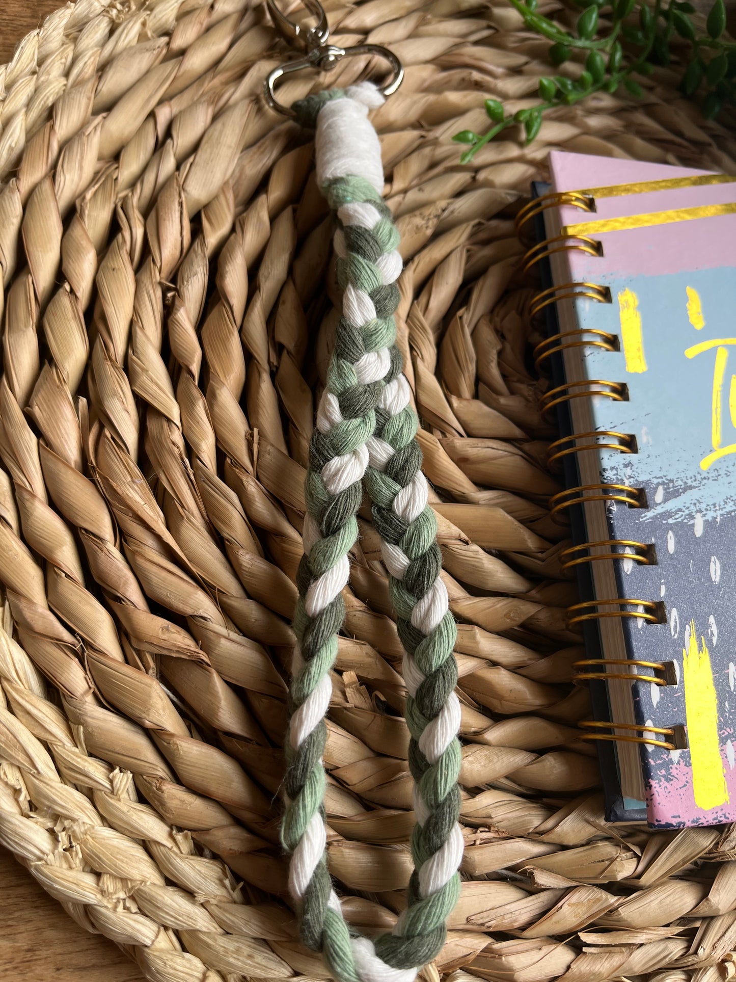 A photo of a handmade macrame braided wristlet, laid on a wicker mat next to a notebook. The wristlet is made with green and natural recycled cotton, with a metal lobster clasp.