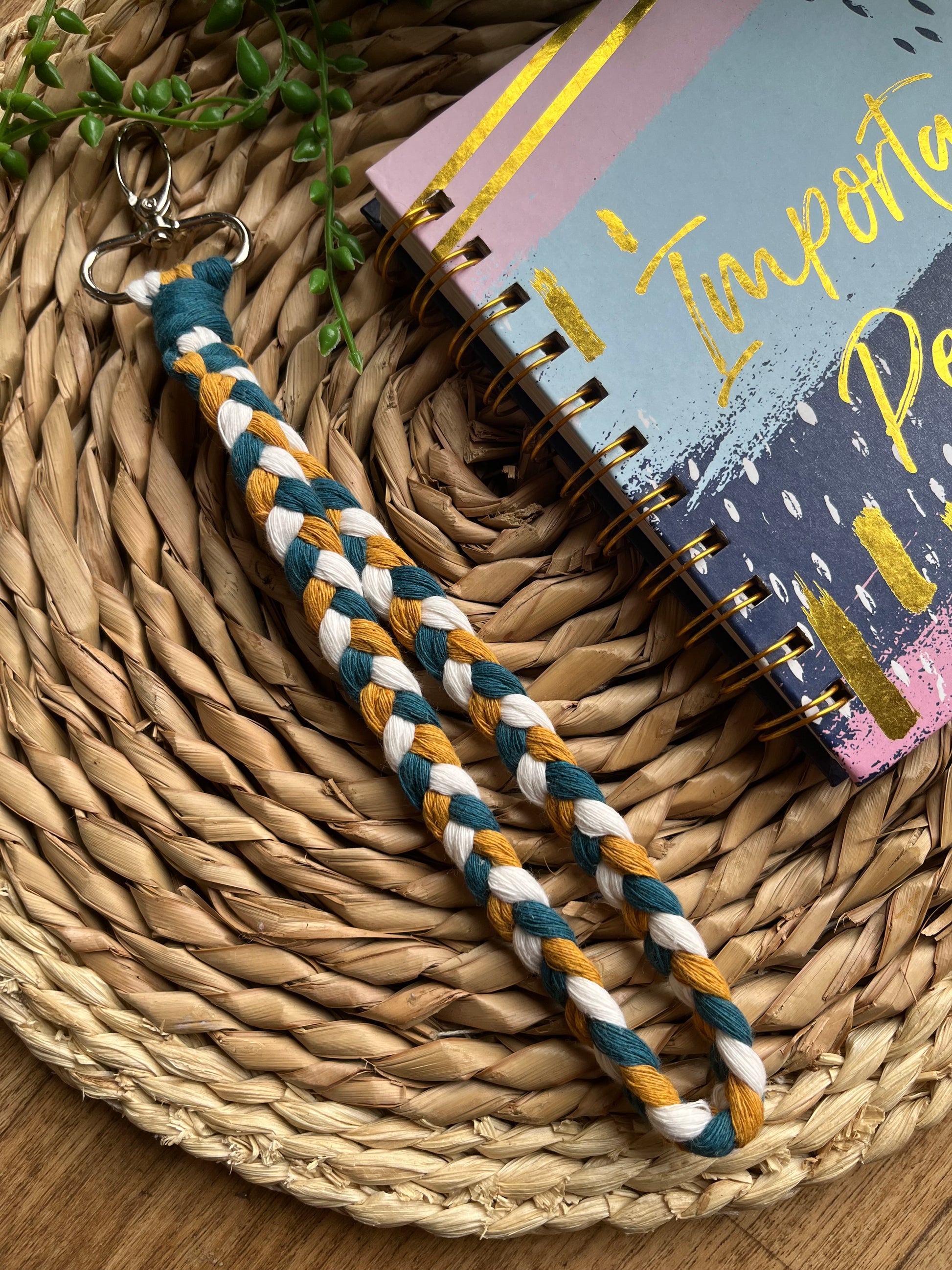 A photo of a handmade macrame braided wristlet, laid on a wicker mat next to a notebook. The wristlet is made with petrol blue, mustard yellow and natural recycled cotton, with a metal lobster clasp.