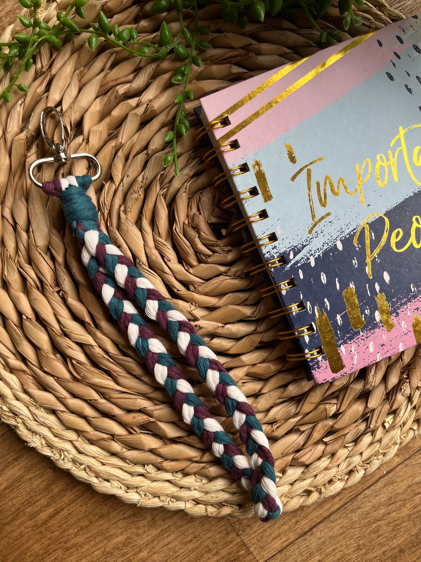 A photo of a handmade macrame braided wristlet, laid on a wicker mat next to a notebook. The wristlet is made with petrol blue, berry purple and natural recycled cotton, with a metal lobster clasp.