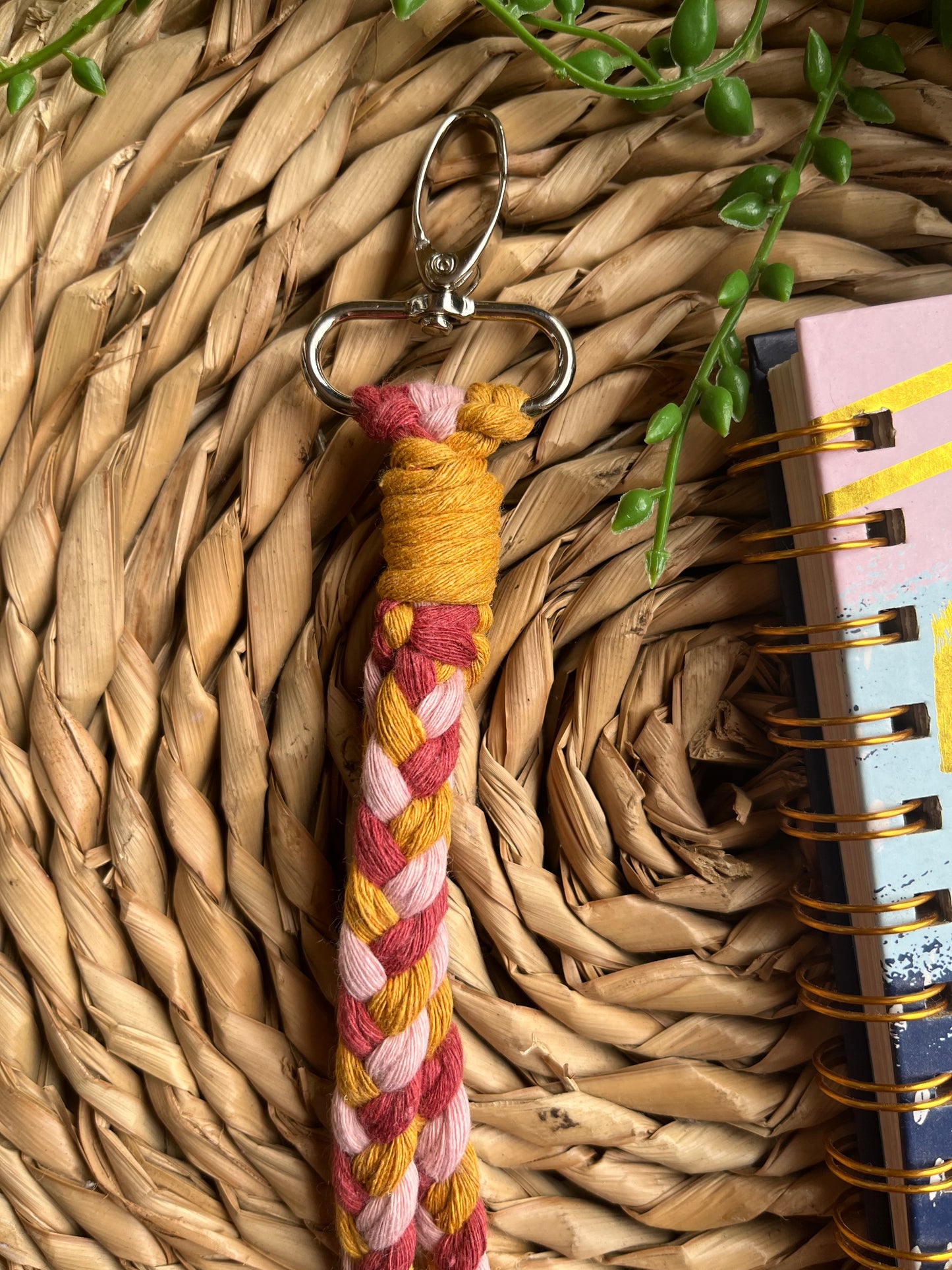 A photo of a handmade macrame braided wristlet, laid on a wicker mat next to a notebook. The wristlet is made with a combination of pink and mustard yellow recycled cottons, with a metal lobster clasp.