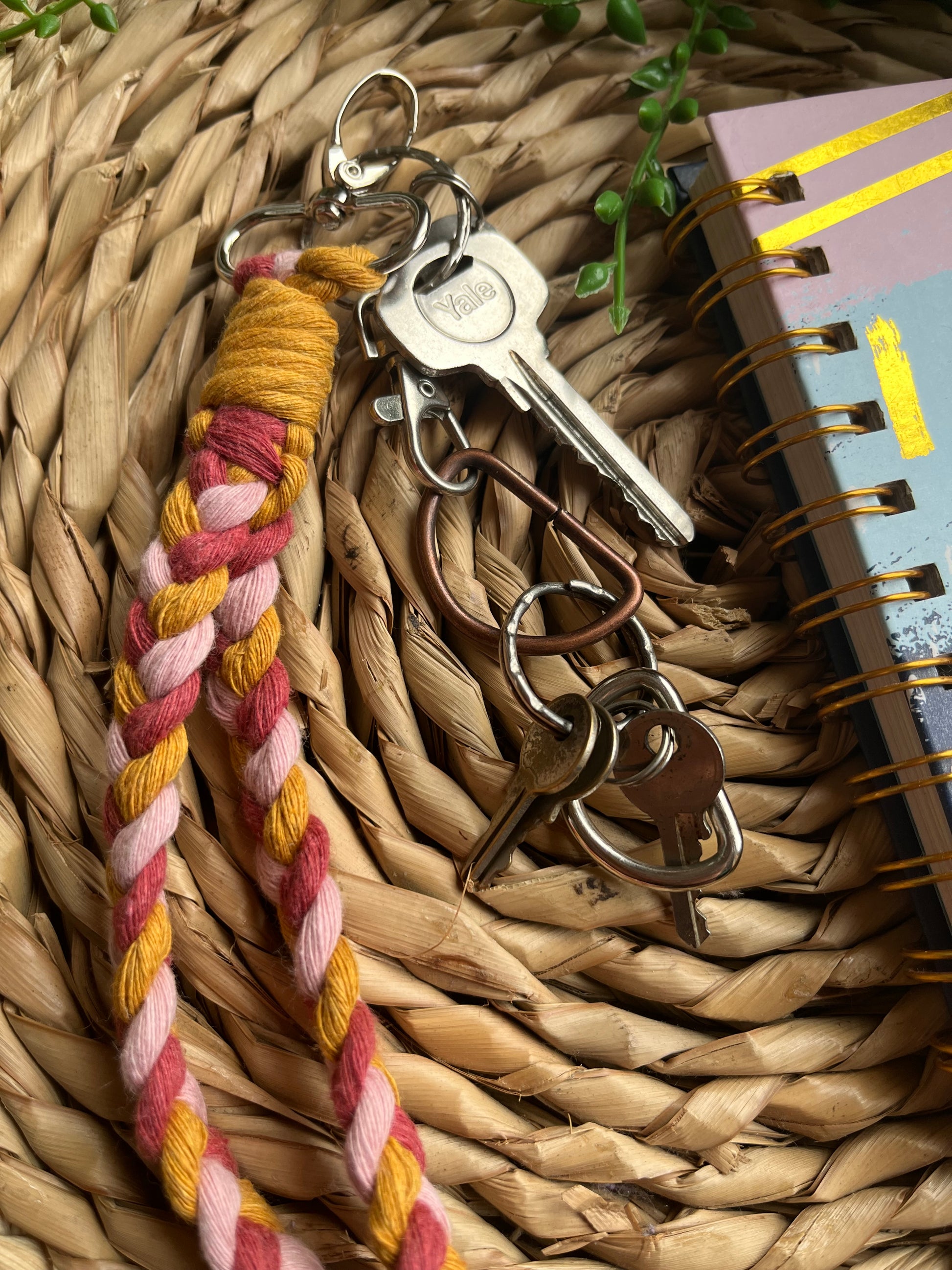 A photo of a handmade macrame braided wristlet attached to a small set of keys, laid on a wicker mat next to a notebook. The wristlet is made with a combination of pink and mustard yellow recycled cottons, with a metal lobster clasp.