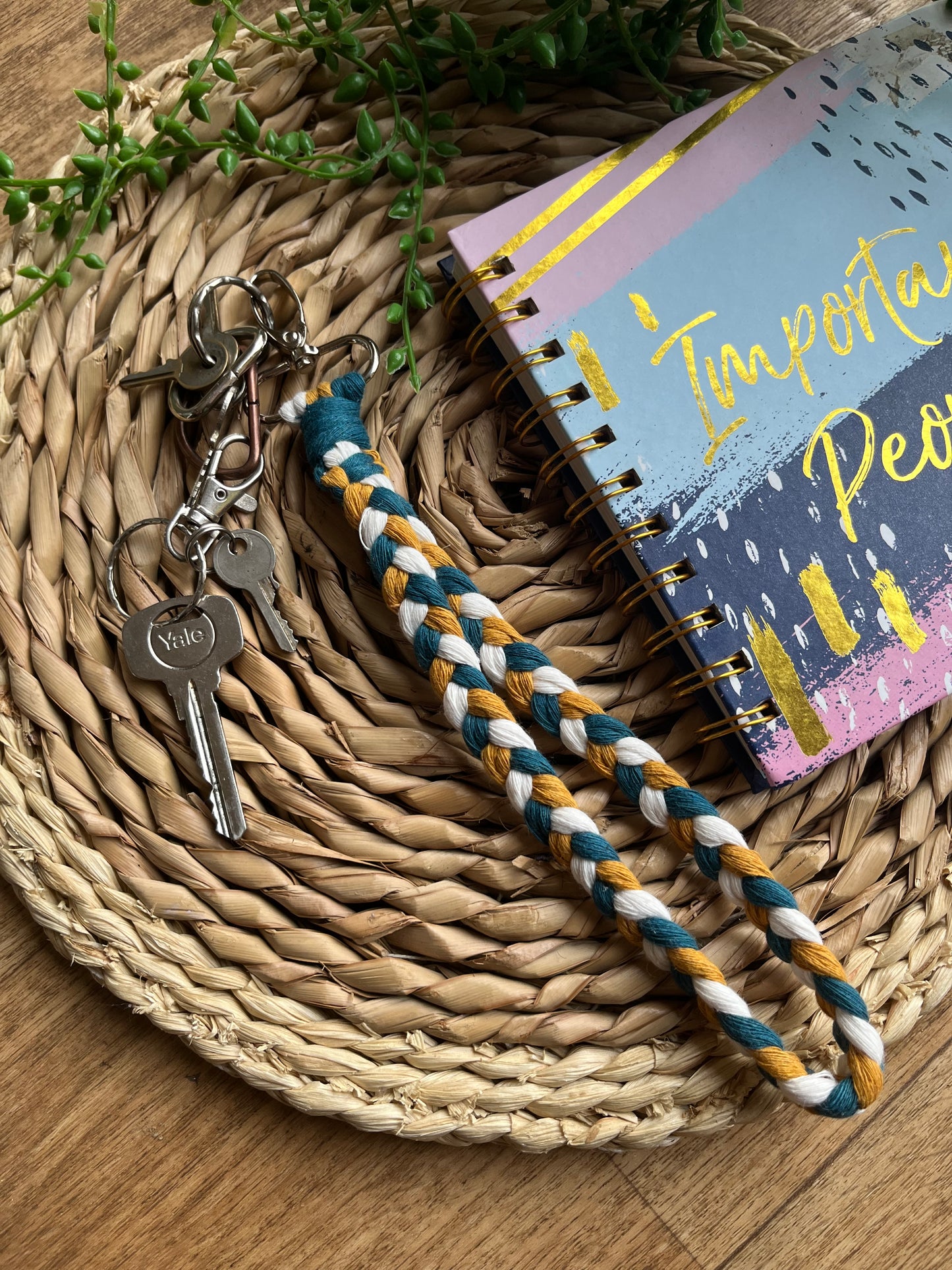 A photo of a handmade macrame braided wristlet attached to a small set of silver keys, laid on a wicker mat next to a notebook. The wristlet is made with petrol blue, mustard yellow and natural recycled cotton, with a metal lobster clasp.