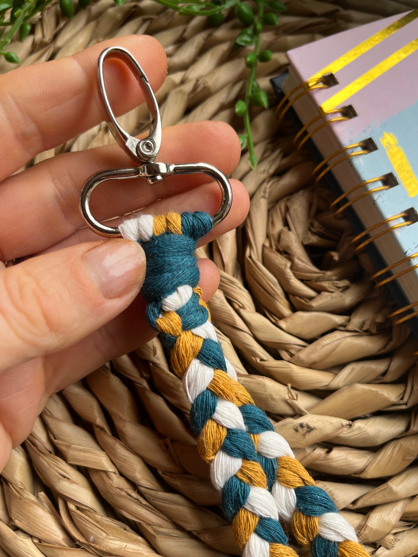 A photo of a hand holding a handmade macrame braided wristlet, on a wicker mat next to a notebook. The wristlet is made with petrol blue, mustard yellow and natural recycled cotton, with a metal lobster clasp.