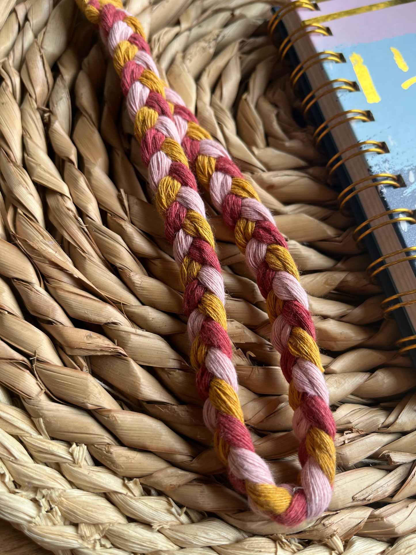 A photo of a handmade macrame braided wristlet, laid on a wicker mat next to a notebook. The wristlet is made with a combination of pink and mustard yellow recycled cottons.