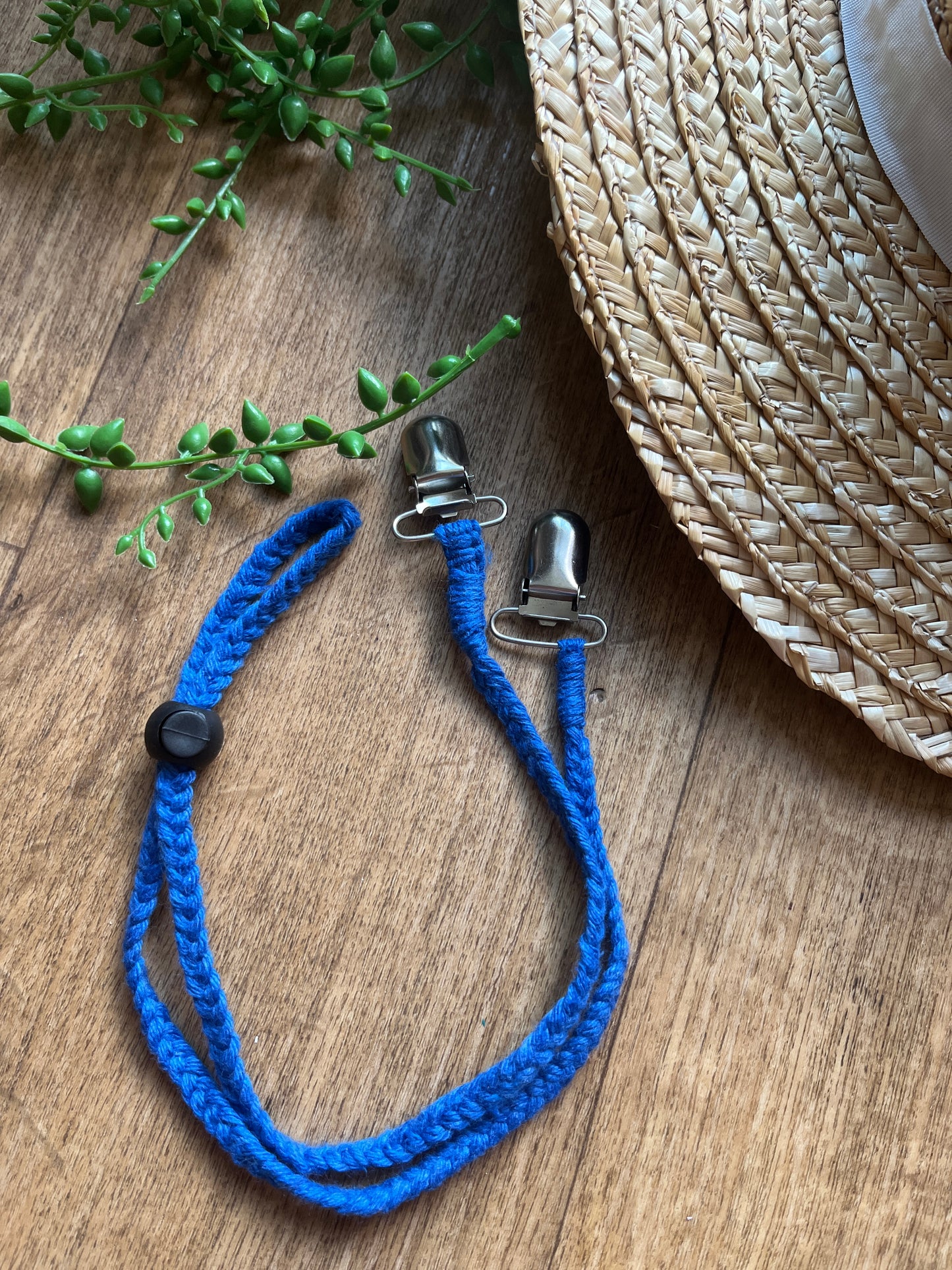 Blue braided macrame chin strap with metal clips and a black toggle on a wooden surface next to a straw hat and plant.