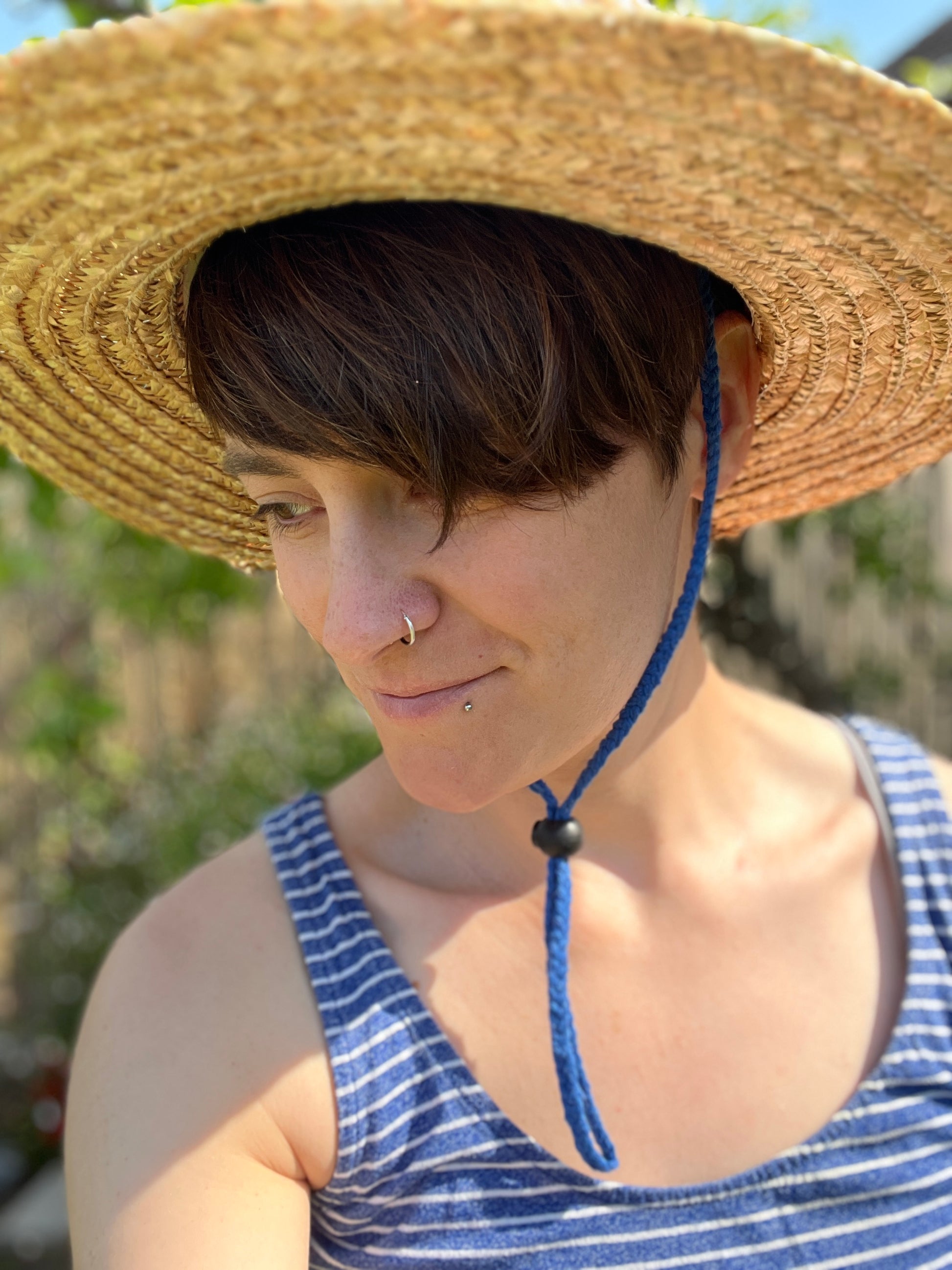 A person with short brown hair wearing a blue striped tank top and straw hat, secured under the chin with blue braided chin strap cord, with a blurred natural background