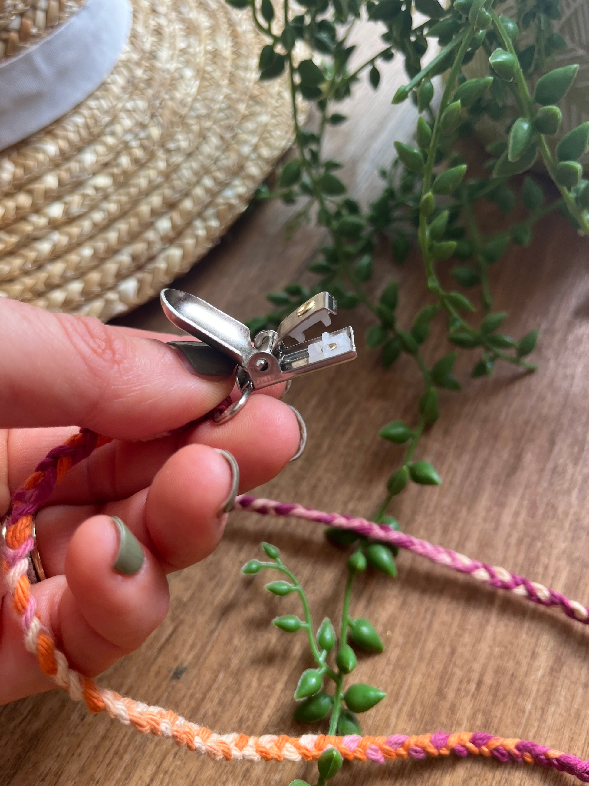 Hand holding a small metal crocodile clasp attached to a braided multicoloured chin strap on a wooden surface with greenery.