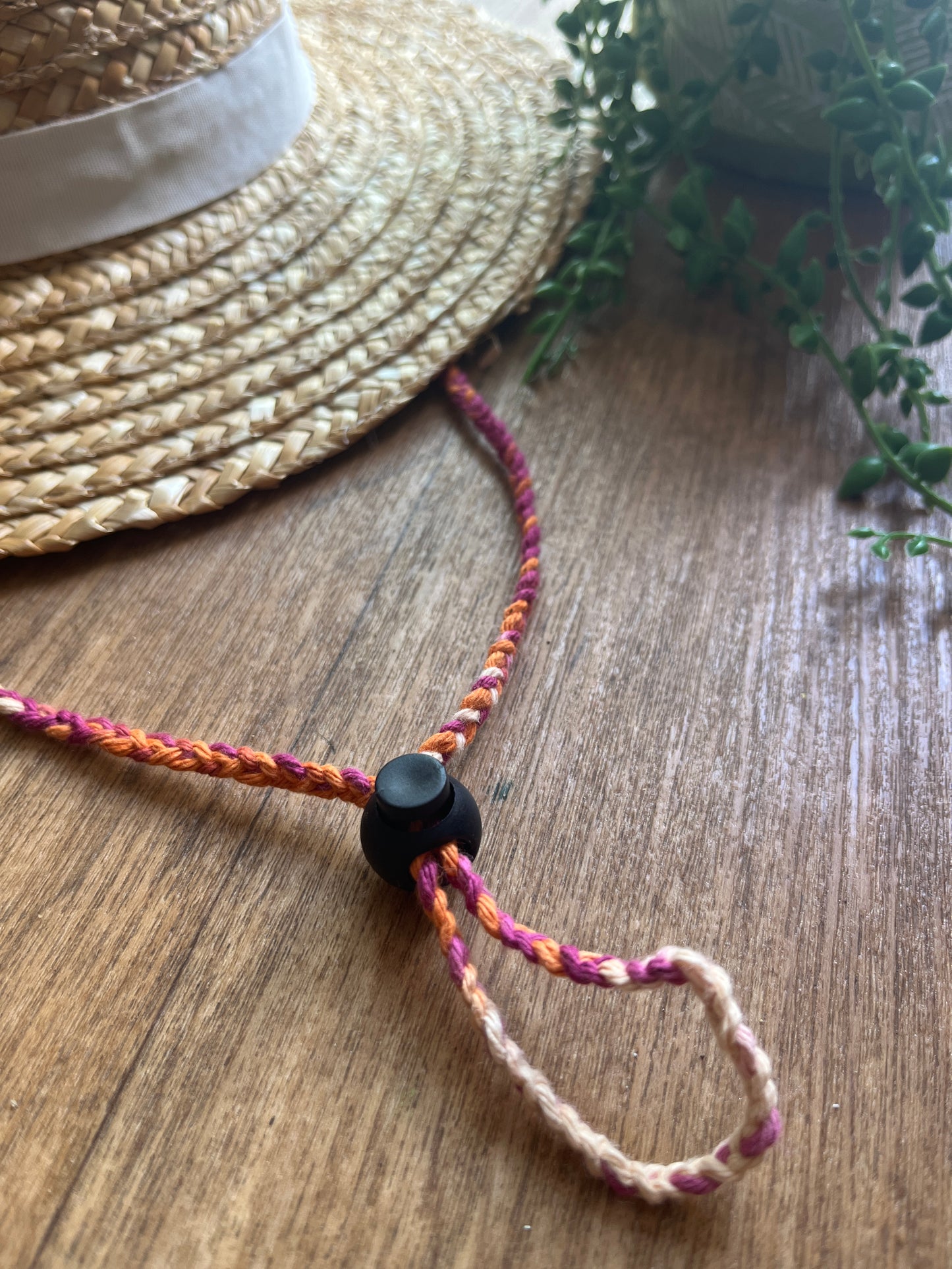 Multicoloured braided chin strap cord with a black toggle on a wooden surface, attached to a straw hat.