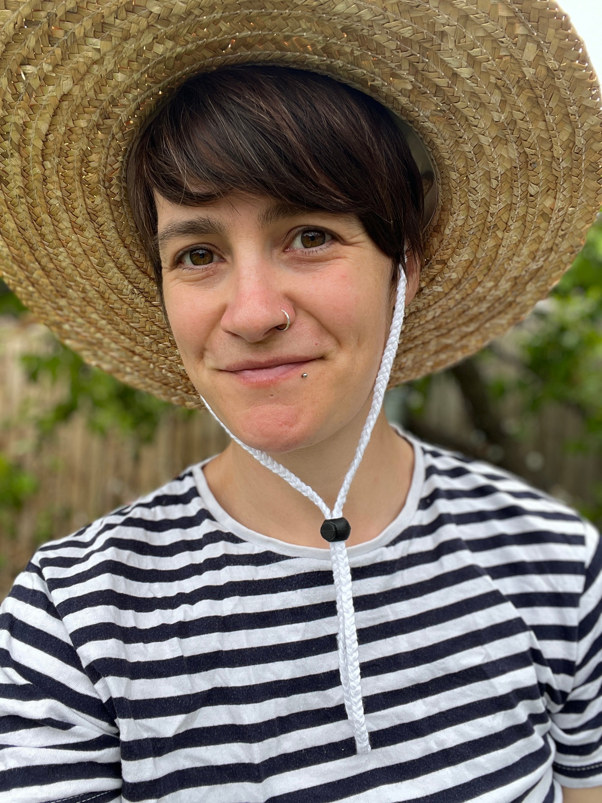 Person wearing a straw hat with white chin secured under the chin with a black toggle, and a striped shirt with a blurred natural background