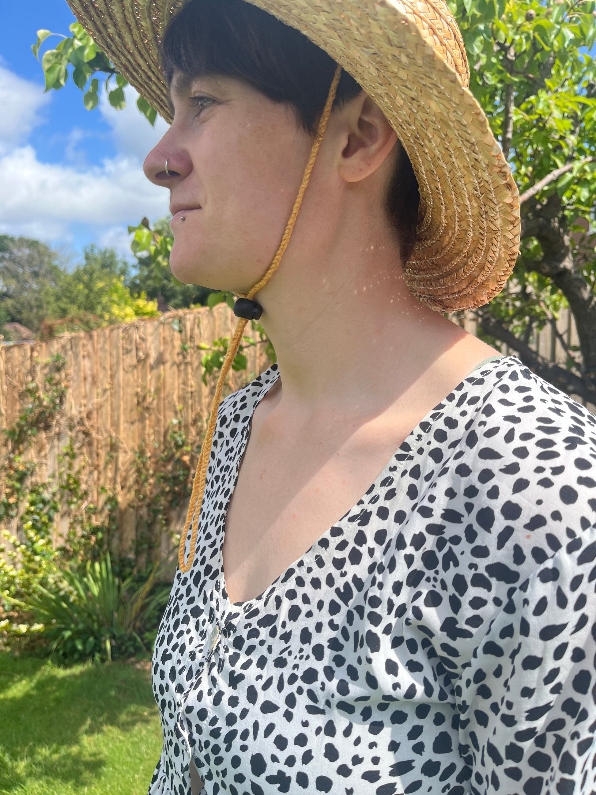A woman with short brown hair wearing a straw hat and patterned dress stands outdoors with trees and sky in the background. The straw hat is secure to their head with a yellow braided chin strap, fastening with a toggle under the chin.