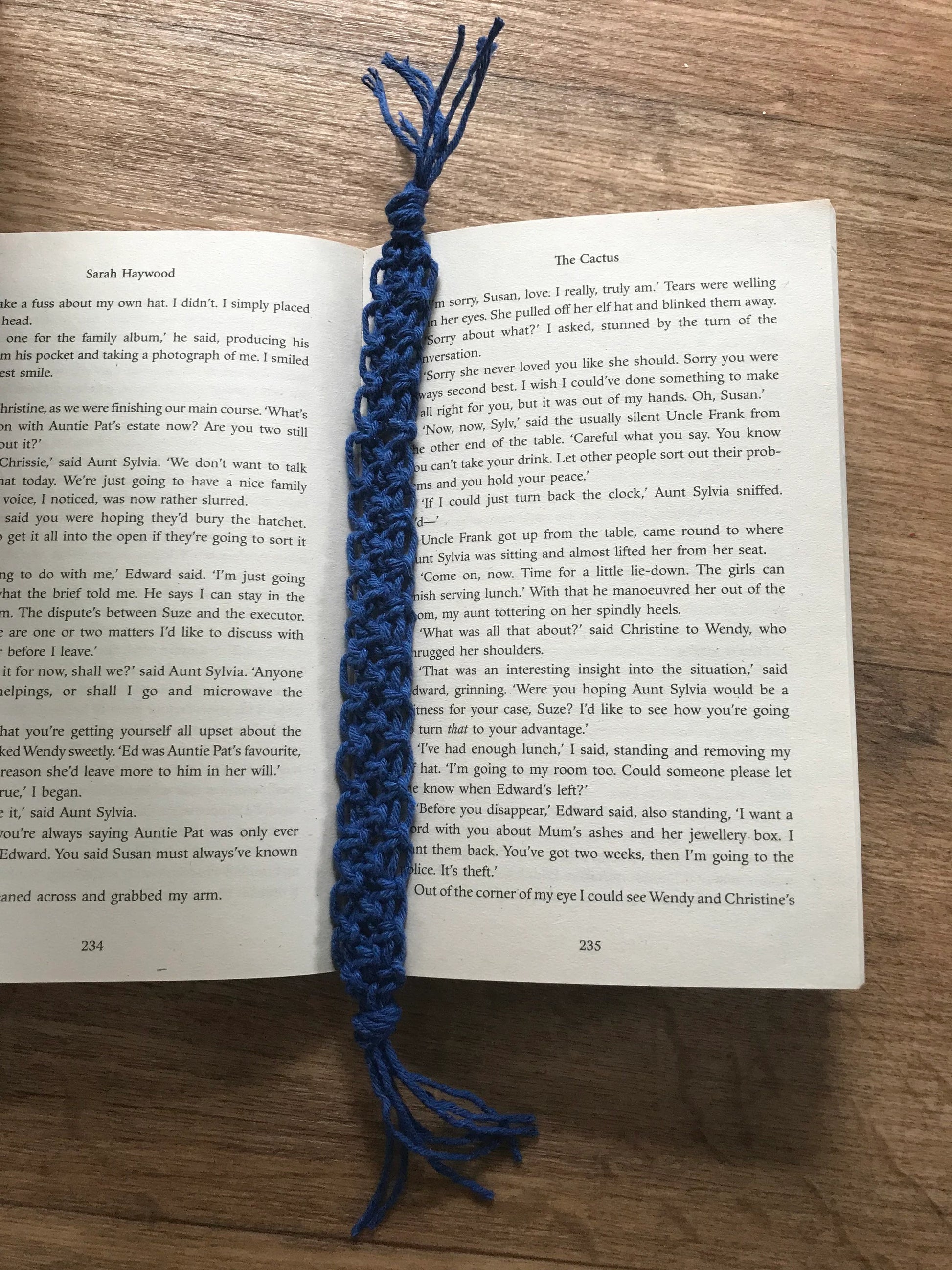 Handmade macramé bookmark with knotted design made with blue recycled cotton, laid across an open book on a wooden surface.