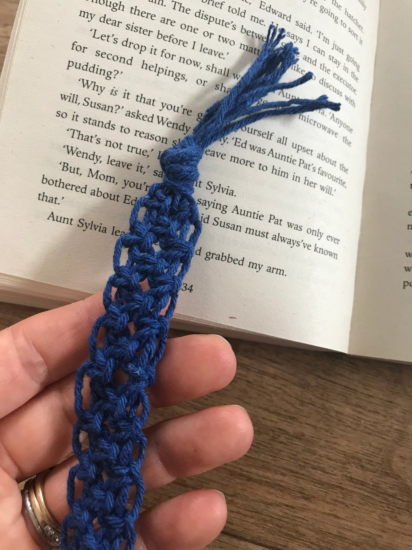 Handmade macramé bookmark with knotted design made with blue recycled cotton, held in a person's hand beside an open book.