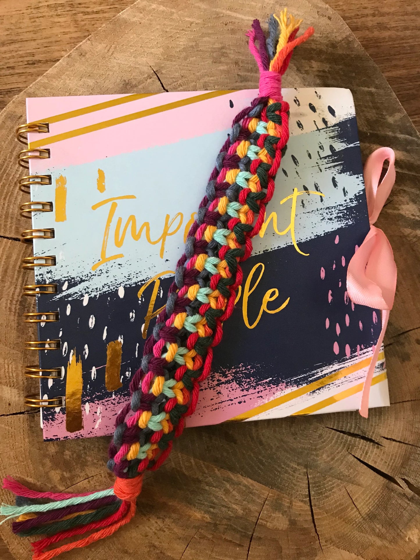 Handmade macramé bookmark with knotted design made with rainbow colours of recycled cotton, laid across a closed address book on a wooden surface.