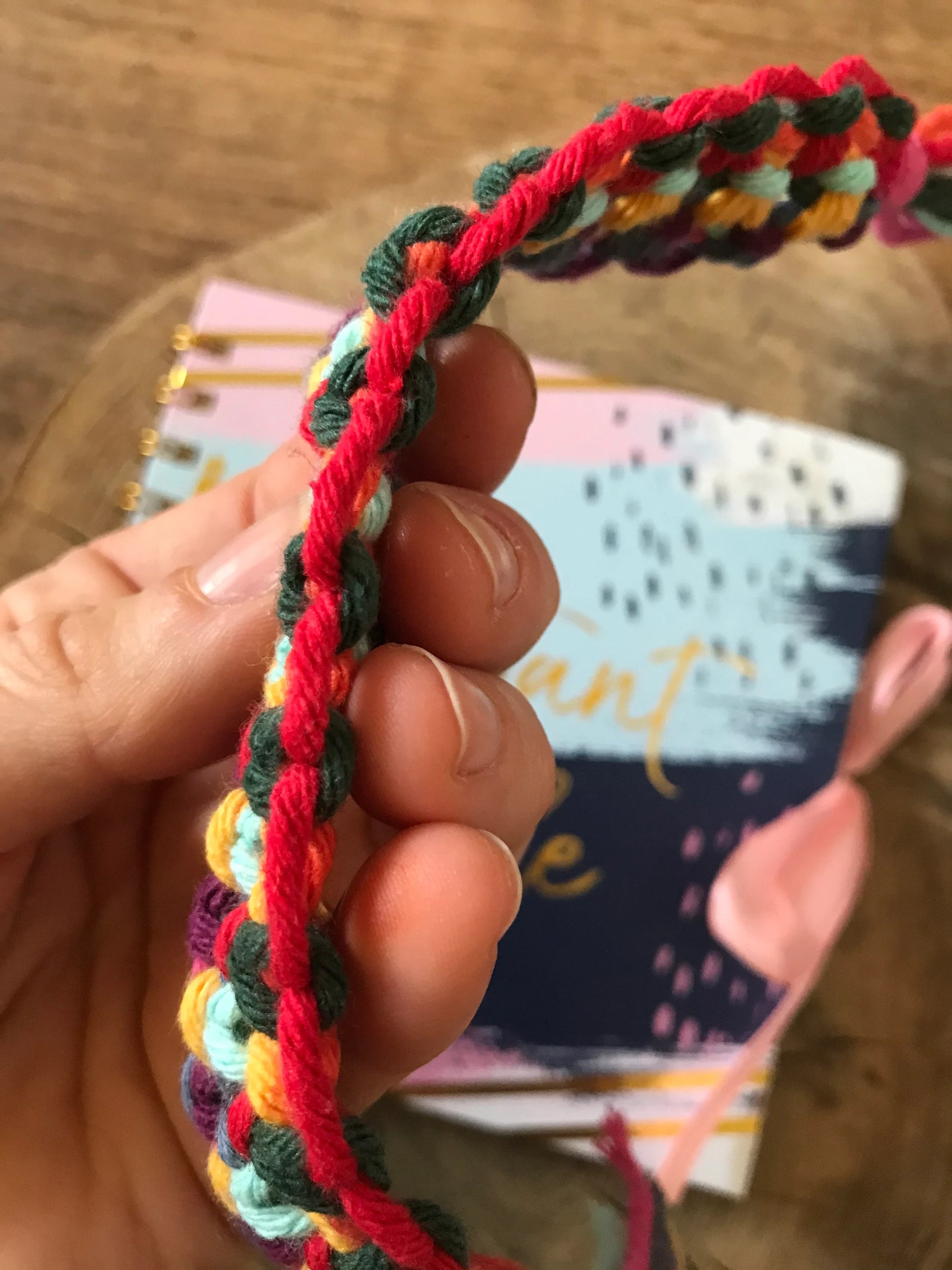 A hand showing a close up of a handmade macramé bookmark with knotted design made with rainbow colours of recycled cotton.