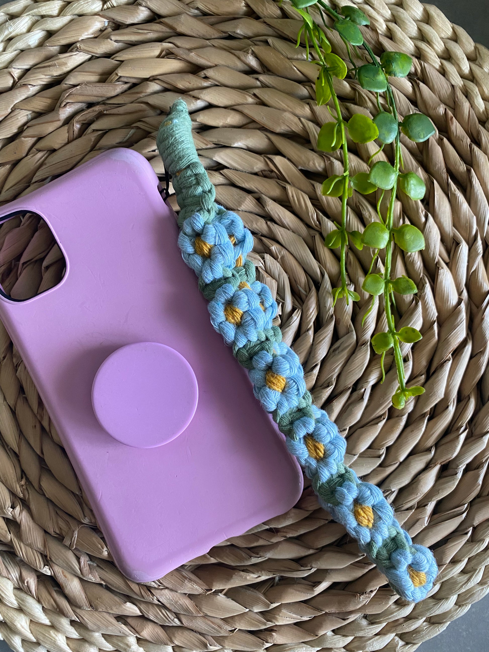 Handmade macrame daisy chain phone charm wristlet, made with blue, yellow and green yarns, attached to a pink phone case with pop socket, and laid on a wicker placemat.