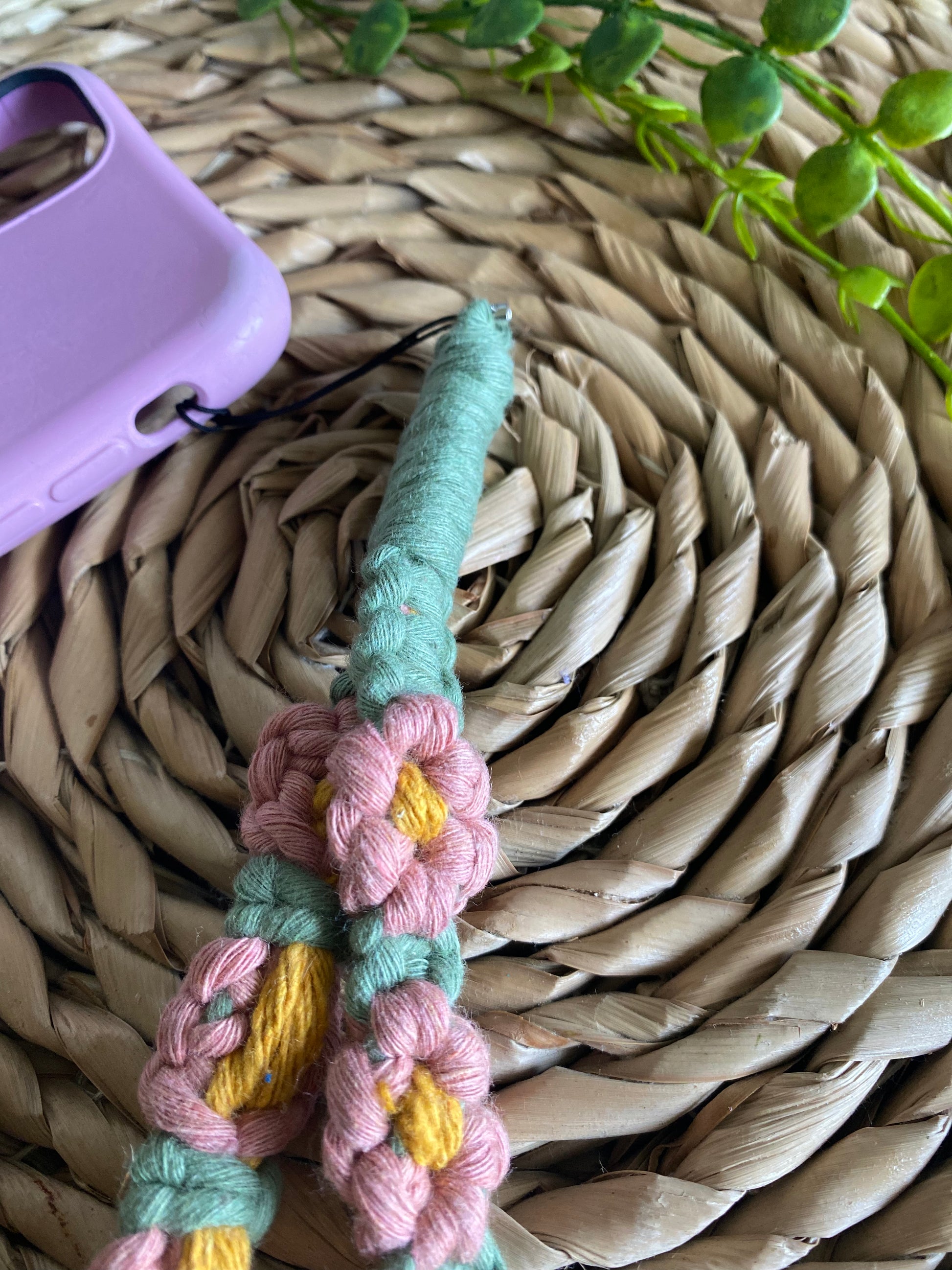 Handmade macrame daisy chain phone charm wristlet, made with pink, yellow and green yarns, attached to a pink phone case, and laid on a wicker placemat.