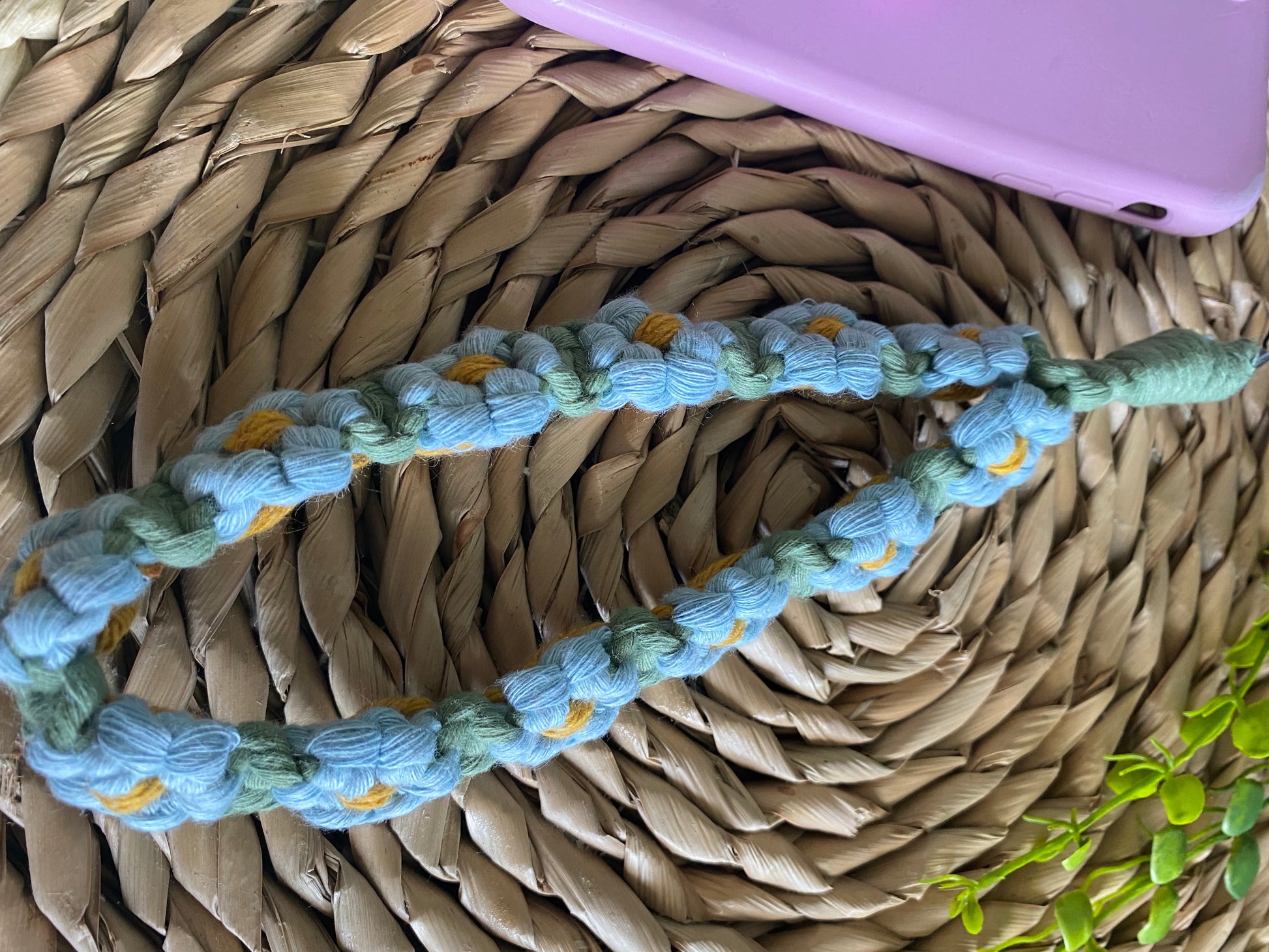 Handmade macrame daisy chain phone charm wristlet, made with blue, yellow and green yarns, attached to a pink phone and laid on a wicker placemat.