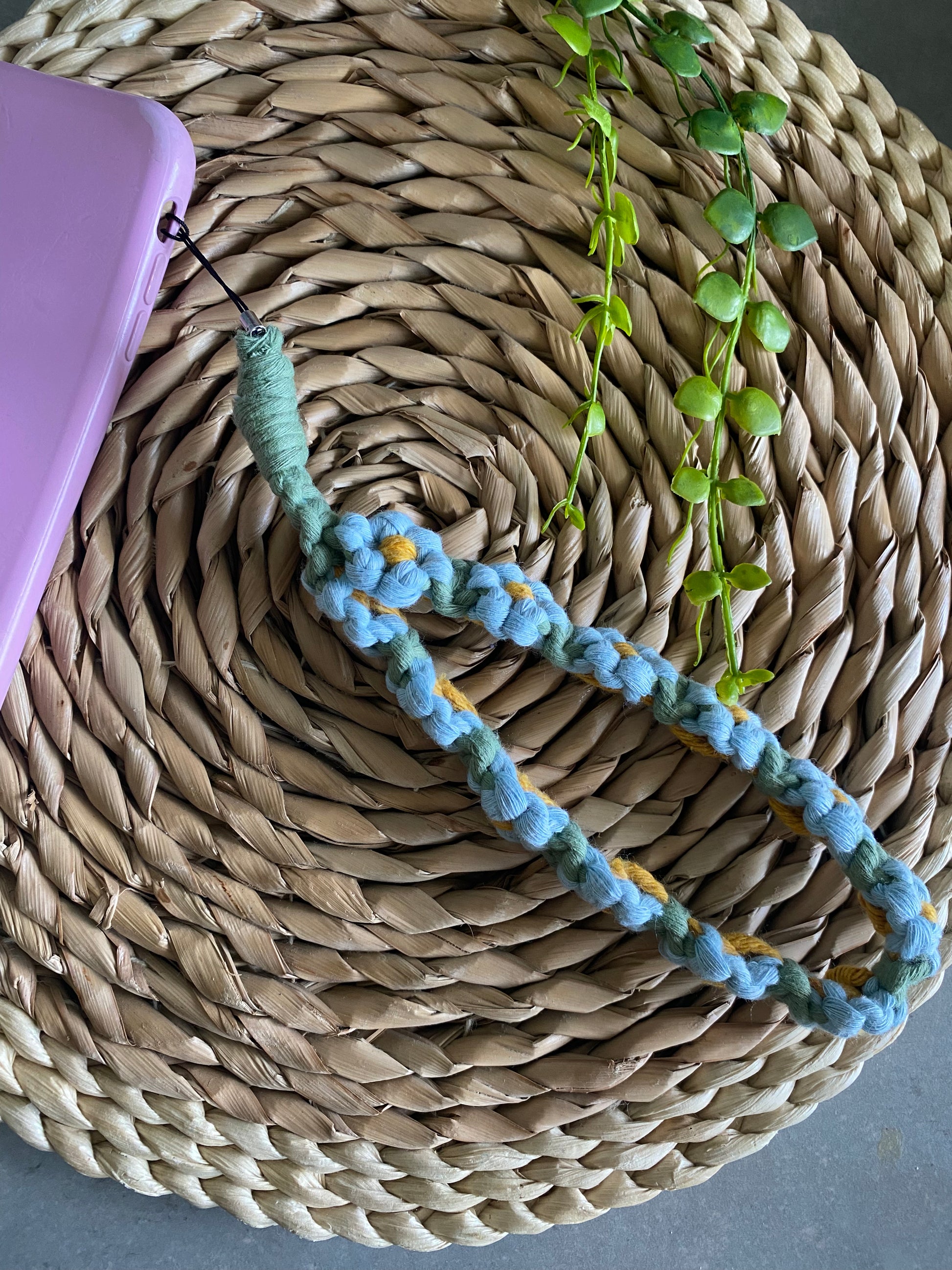 Close up of a macrame daisy chain phone charm wristlet, made with blue, yellow and green yarns, attached to a pink phone case, and laid on a wicker placemat.