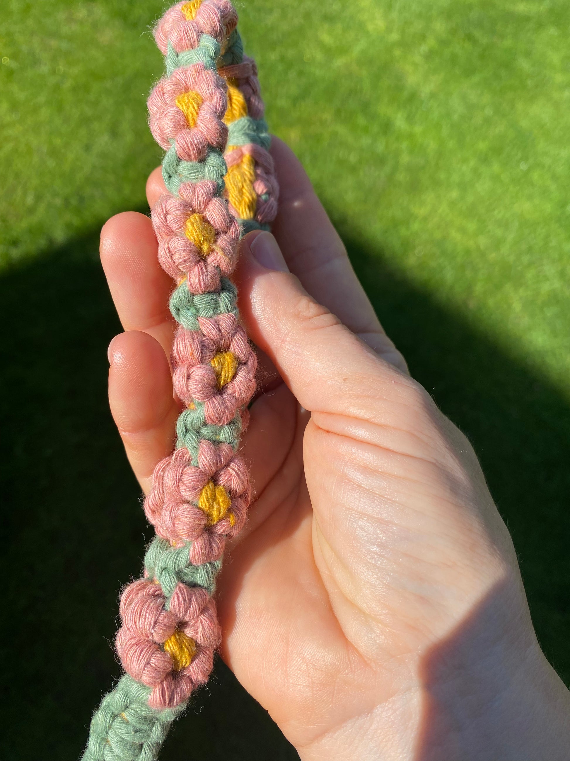 A hand holding a macrame daisy chain phone charm wristlet, made with pink, yellow and green yarns with lush green grass in the background.