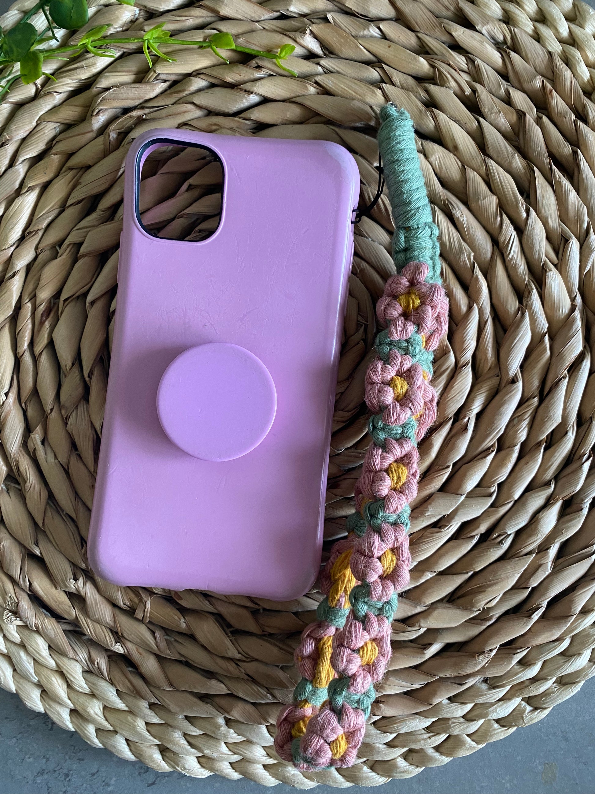 Handmade macrame daisy chain phone charm wristlet, made with pink, yellow and green yarns, attached to a pink phone case with pop socket, and laid on a wicker placemat.