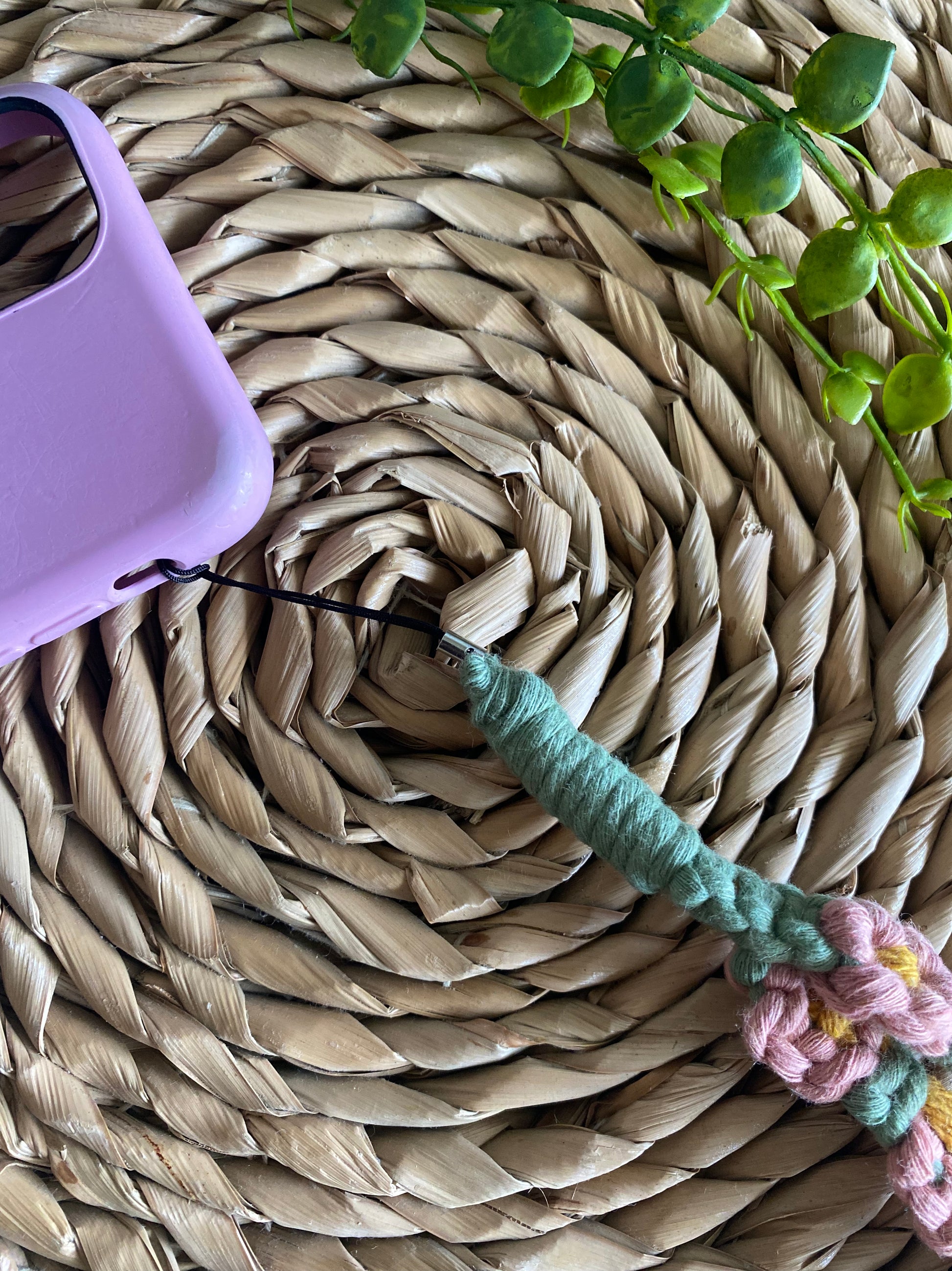 Close up of a macrame daisy chain phone charm wristlet, made with pink, yellow and green yarns, attached to a pink phone case and laid on a wicker placemat.