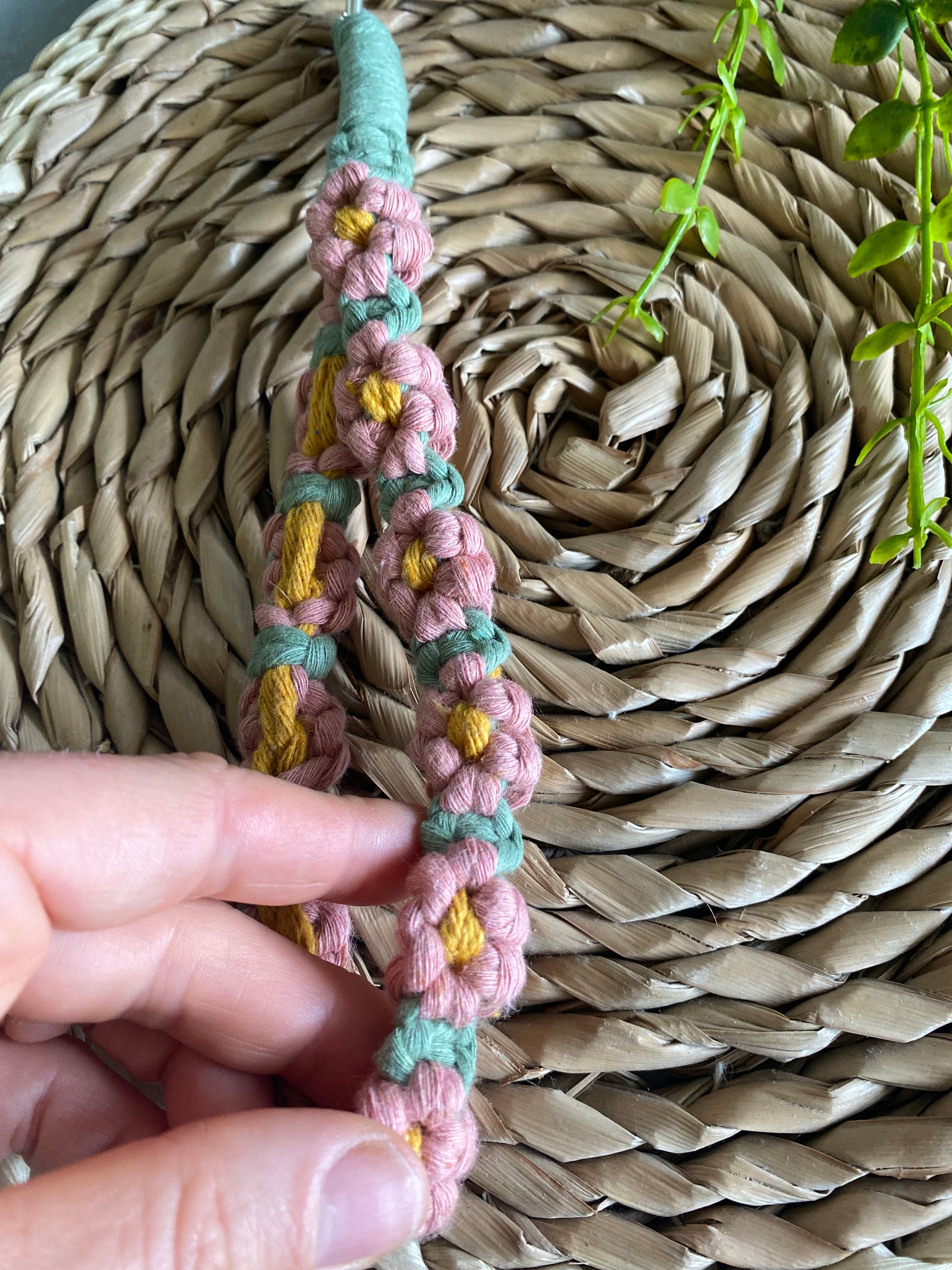 A hand showing a macrame daisy chain phone charm wristlet, made with pink, yellow and green yarns.