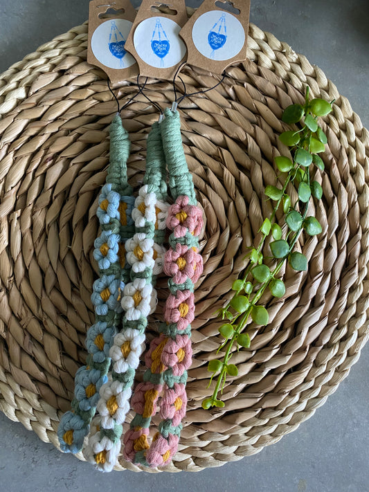 Three macrame daisy chain phone charm wristlets, made in different colours, each attached to a cardboard swing tag, and laid next to each other on a wicker placemat.