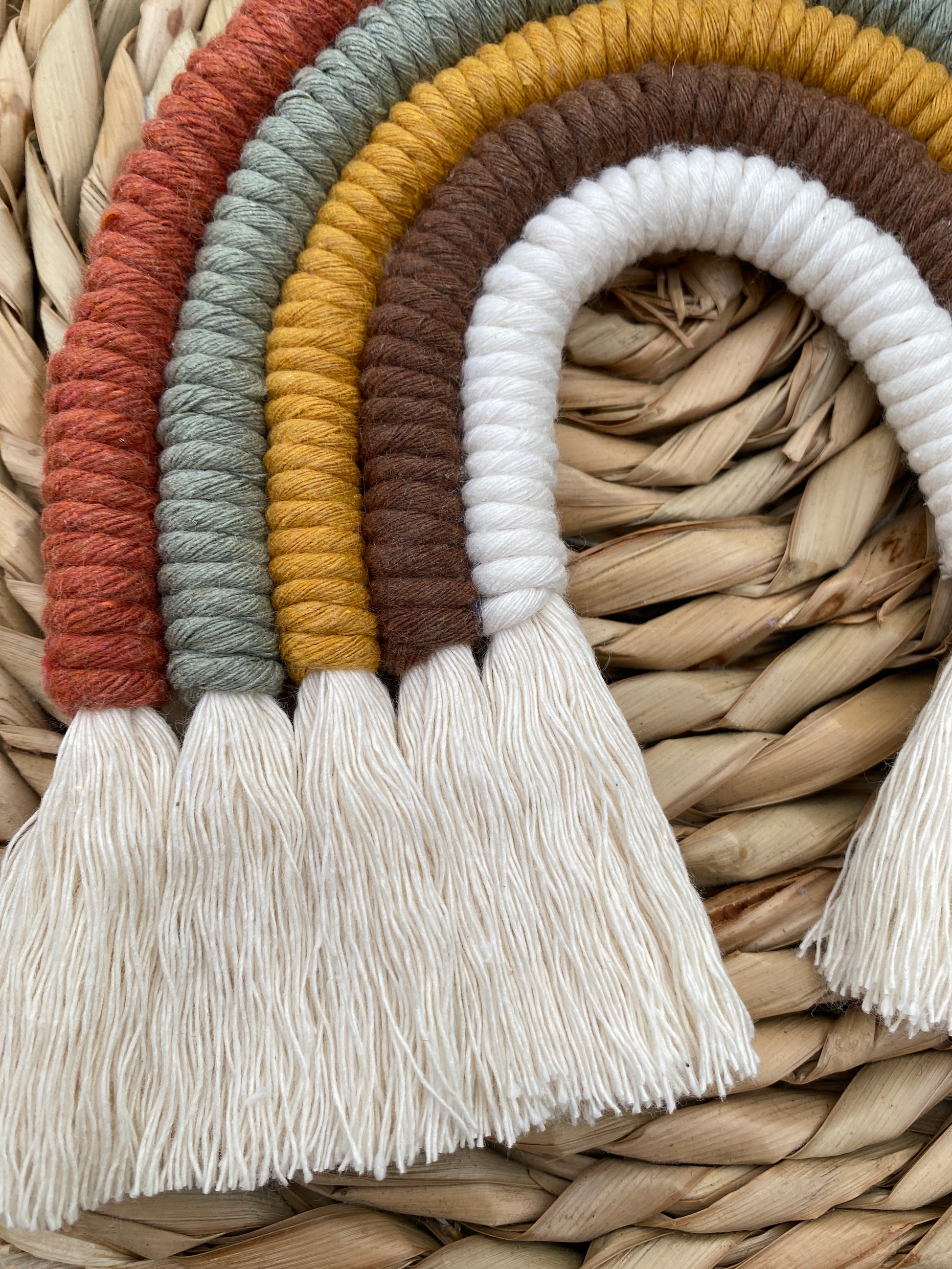 Close up of a handmade macrame rainbow, in earthy brown, mustard, green tones, with natural fringed ends, placed on a wicker placemat.