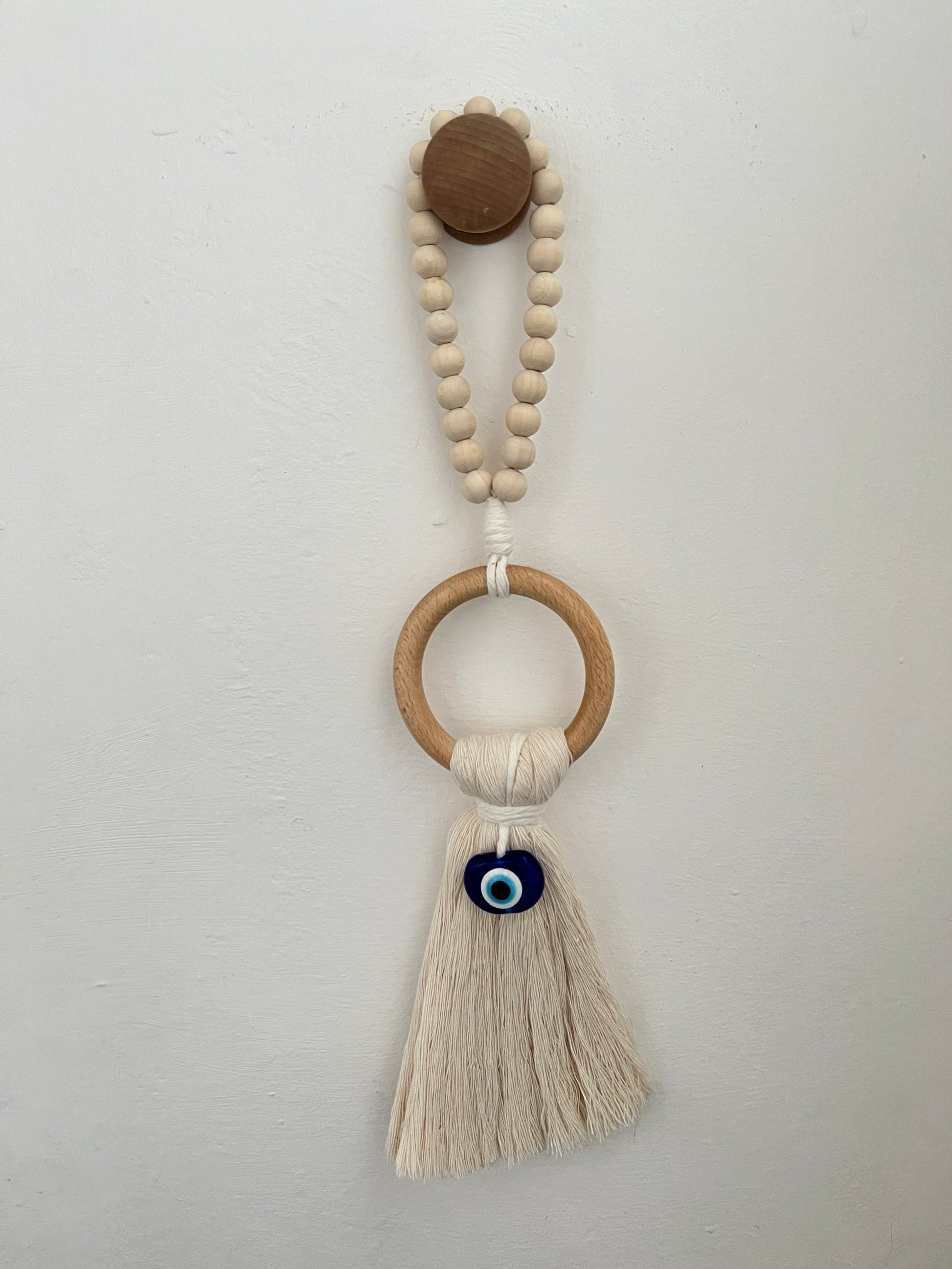 Handmade macrame door hanger, made with recycled cotton, on a wooden ring, with a blue resin evil eye bead and beaded hanging loop. Shown hanging from a wooden knob on a white wall.