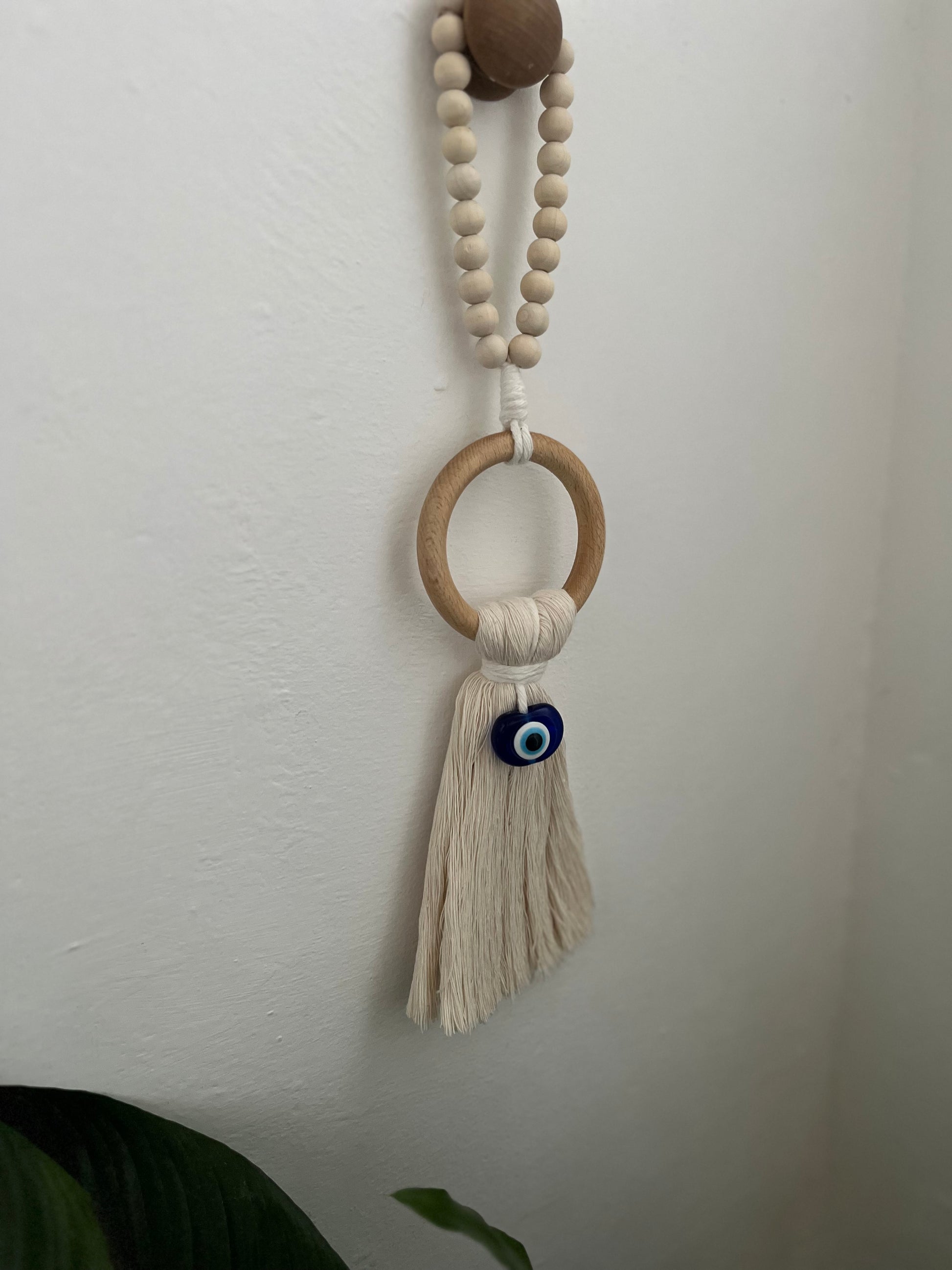 Handmade macrame door hanger, made with recycled cotton, on a wooden ring, with a blue resin evil eye bead and a beaded hanging loop. Hanging from a wooden knob on a white wall.