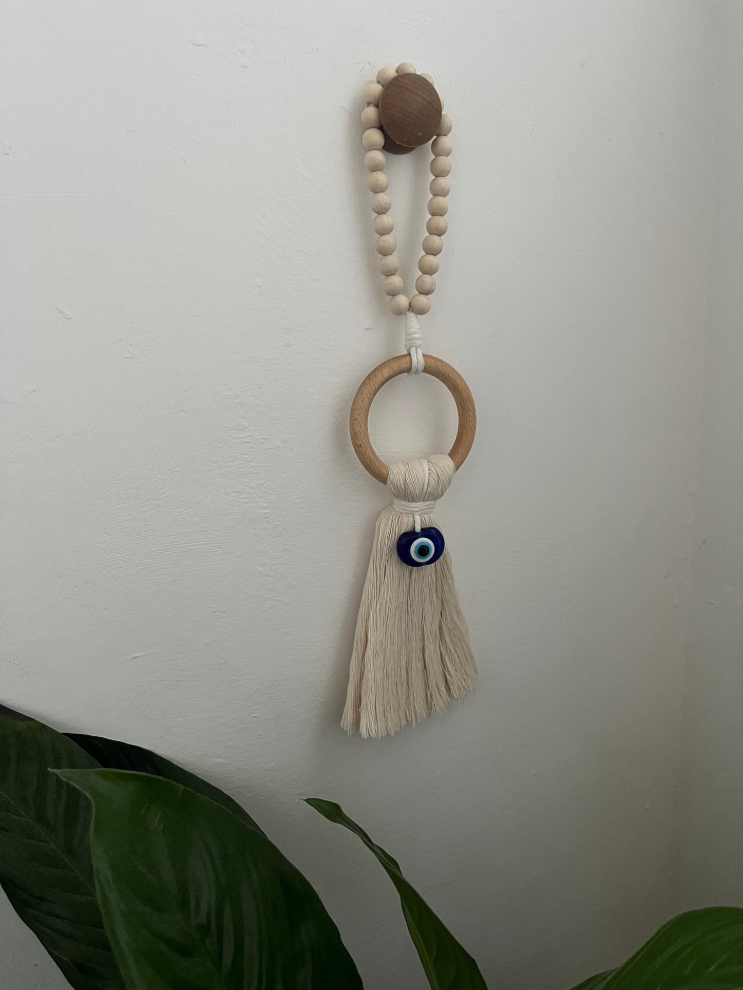Handmade macrame door hanger, made with recycled cotton, on a wooden ring, with a blue resin evil eye bead and beaded hanging loop. Shown hanging from a wooden knob on a white wall.