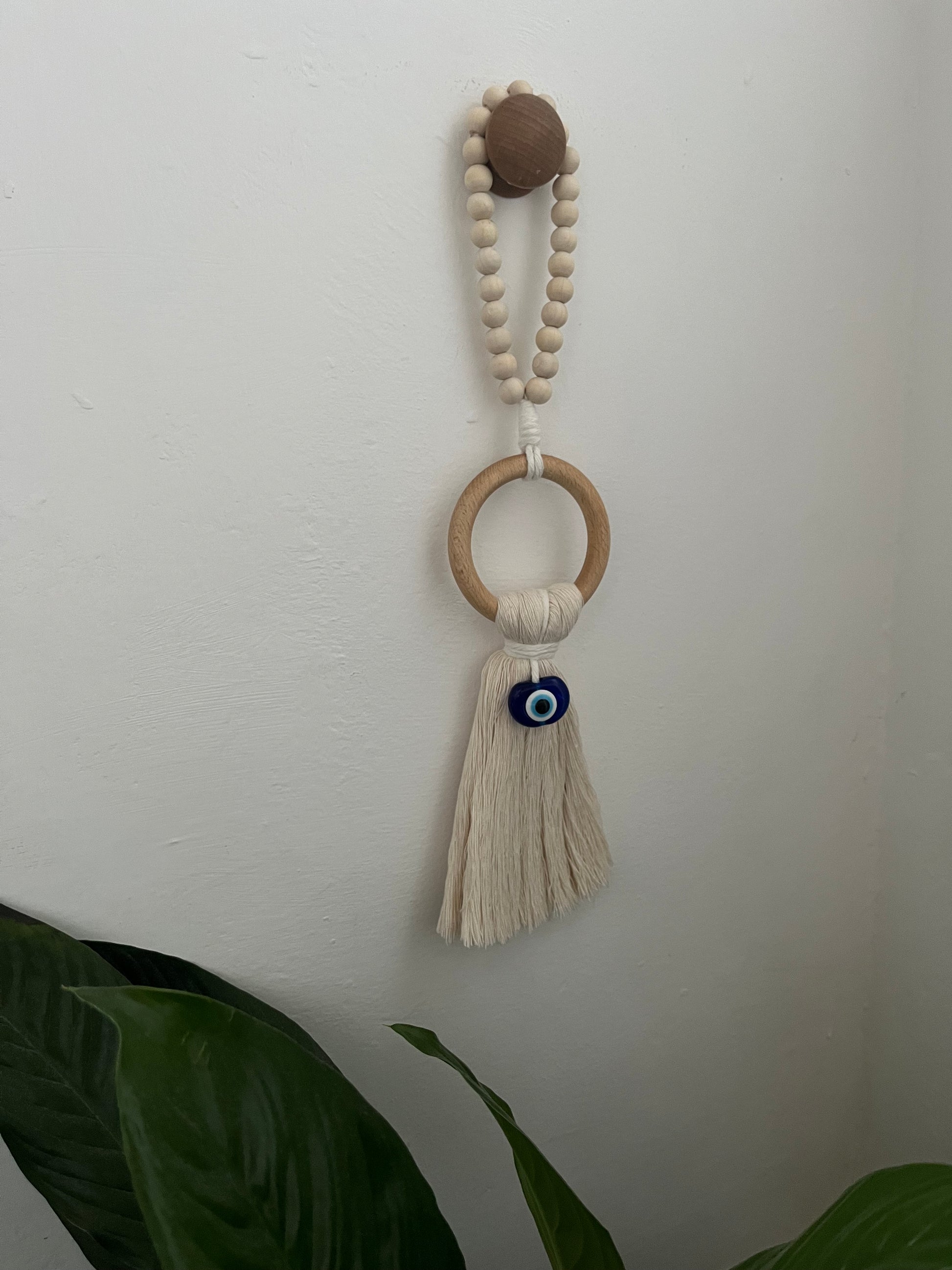 Handmade macrame door hanger, made with recycled cotton, on a wooden ring, with a blue resin evil eye bead and beaded hanging loop. Shown hanging from a wooden knob on a white wall.