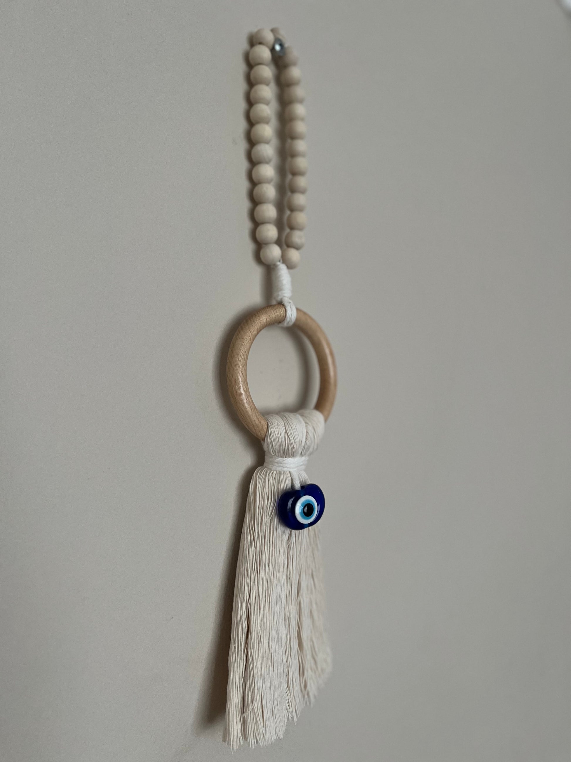 Handmade macrame door hanger, made with recycled cotton, on a wooden ring, with a blue resin evil eye bead and beaded hanging loop. Shown hanging from a screw on a white wall.