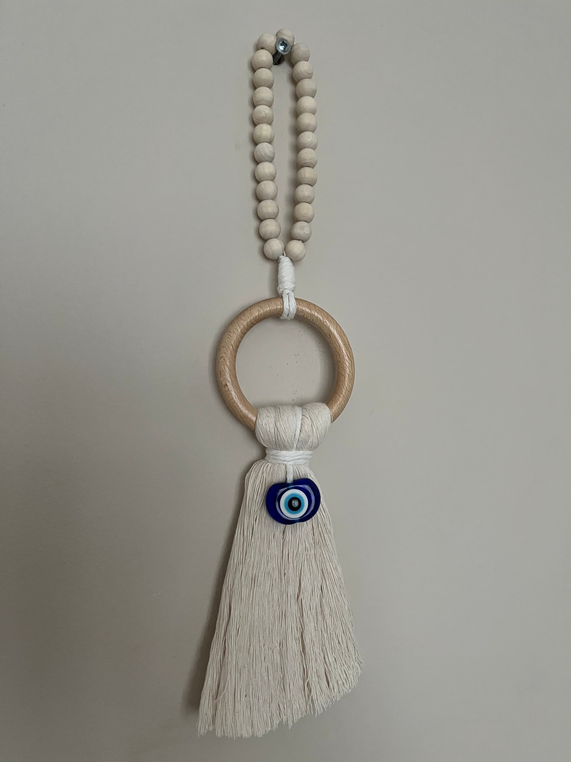 Handmade macrame door hanger, made with recycled cotton, on a wooden ring, with a blue resin evil eye bead and beaded hanging loop. Shown hanging from a screw on a white wall.