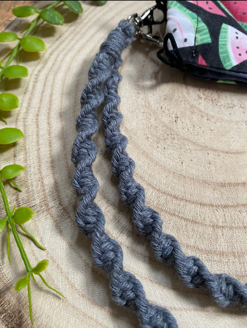 Close up of a handmade macramé face mask lanyard in dark grey cotton cord attached by small metal clips on to a face mask which has a watermelon print.