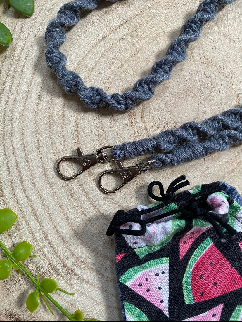 Close up of a handmade macramé face mask lanyard in dark grey cotton cord with small metal clips on each end, placed on a wooden surface next to a face mask which has a watermelon print.