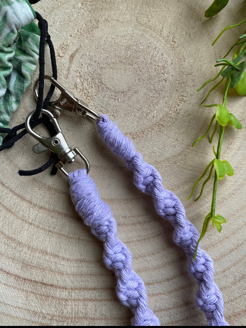 Close up of a handmade macramé face mask lanyard in lilac cotton cord attached by small metal clips on to a face mask which has a green leafy print.