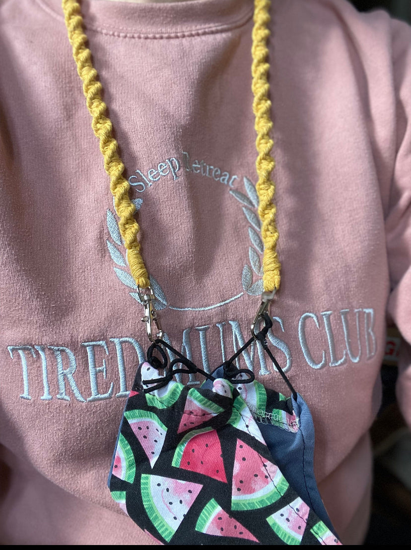 A woman wearing a pink jumper with a handmade macramé face mask lanyard around her neck, attached by small metal clips on to a face mask which has a watermelon print.