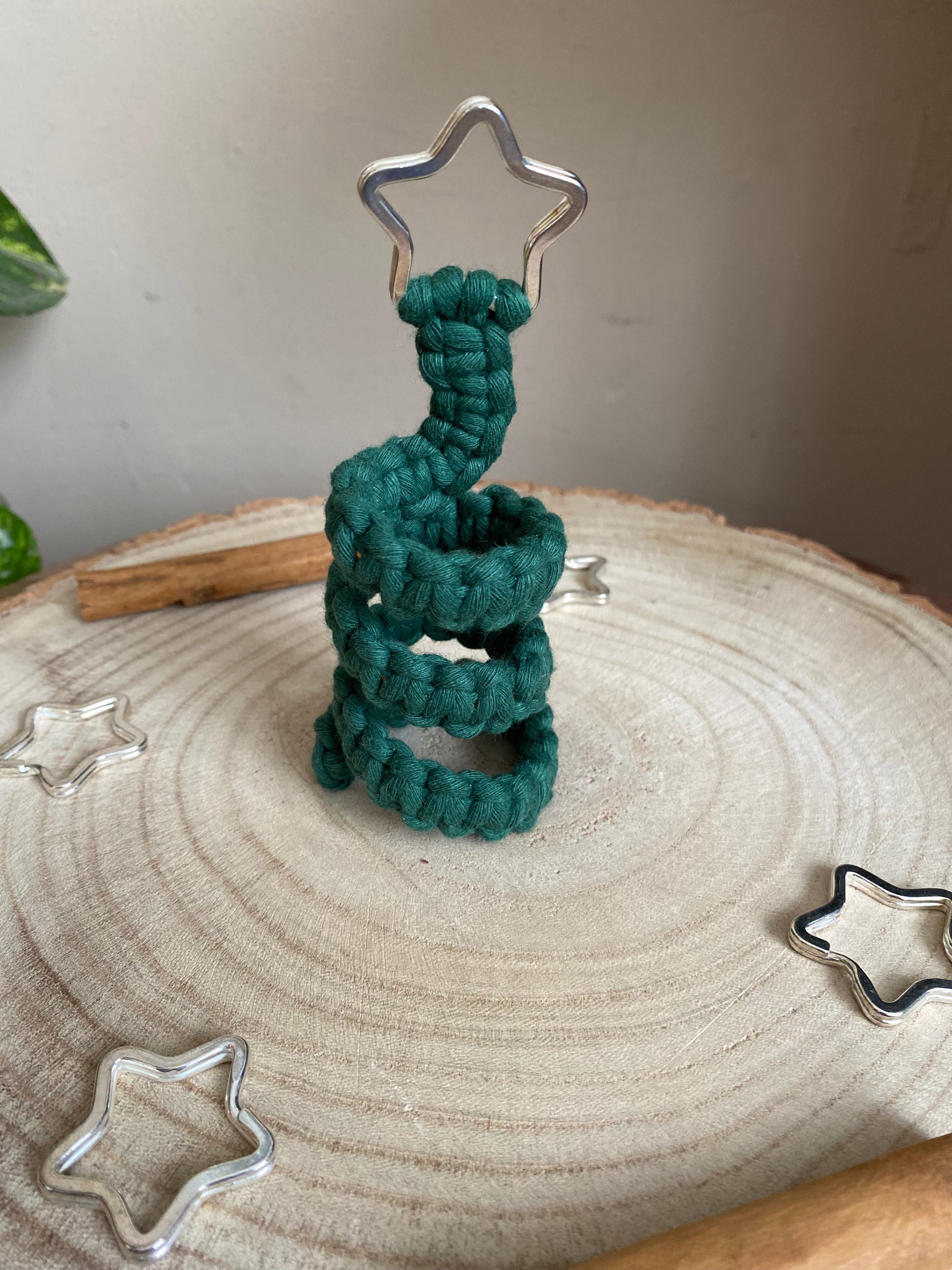 A freestanding spiral Christmas tree ornament made with macrame knots in green yarn with a silver star on top, displayed on a wooden surface with metal stars and cinnamon sticks for decoration.