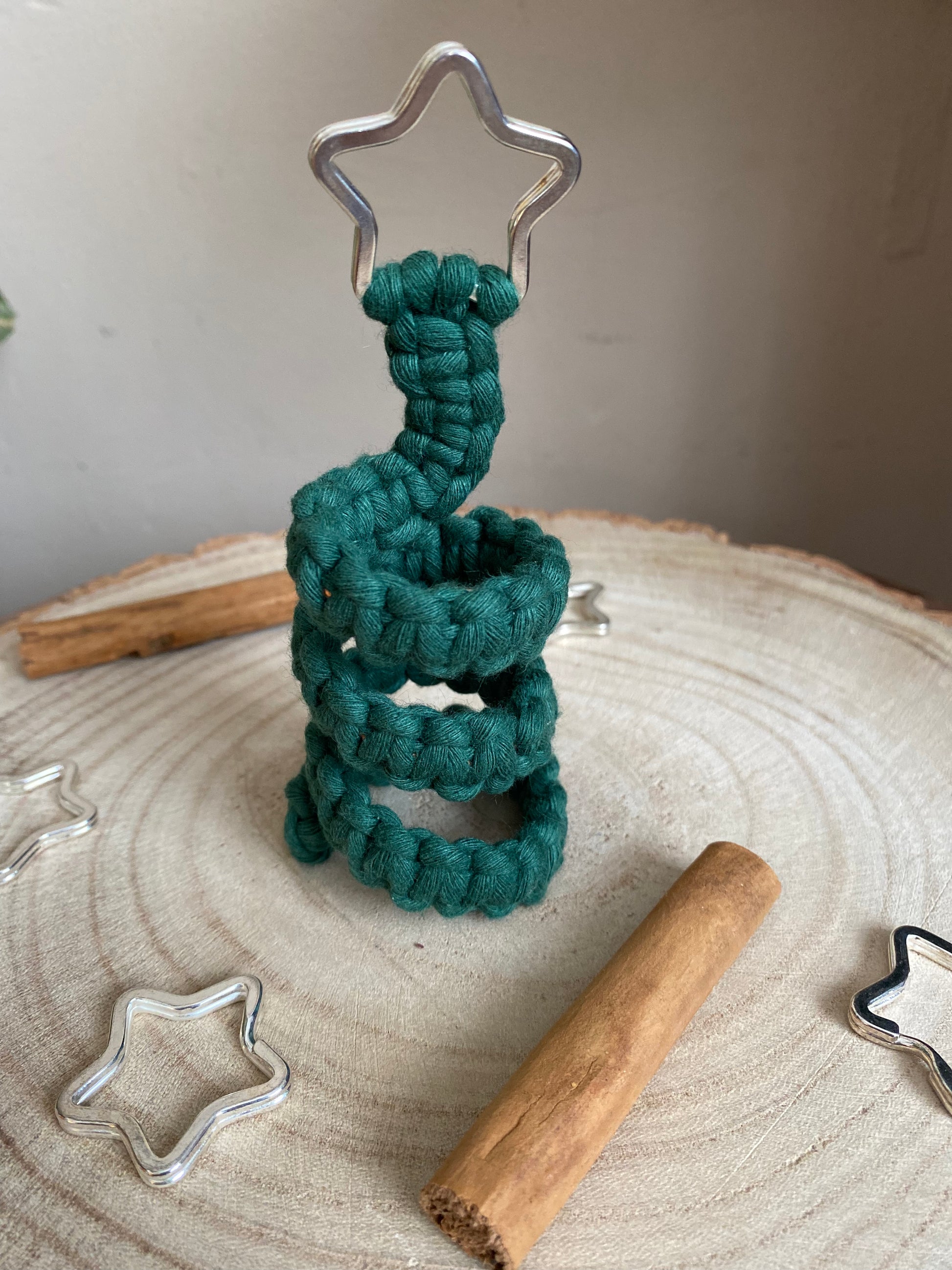 A freestanding spiral Christmas tree ornament made with macrame knots in green yarn with a silver star on top, displayed on a wooden surface with metal stars and cinnamon sticks for decoration.